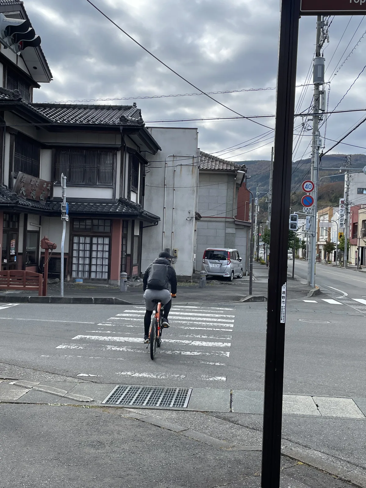 遠野市街的街角，沿街為兩層木造町家，路口有行人與自行車，天空多雲