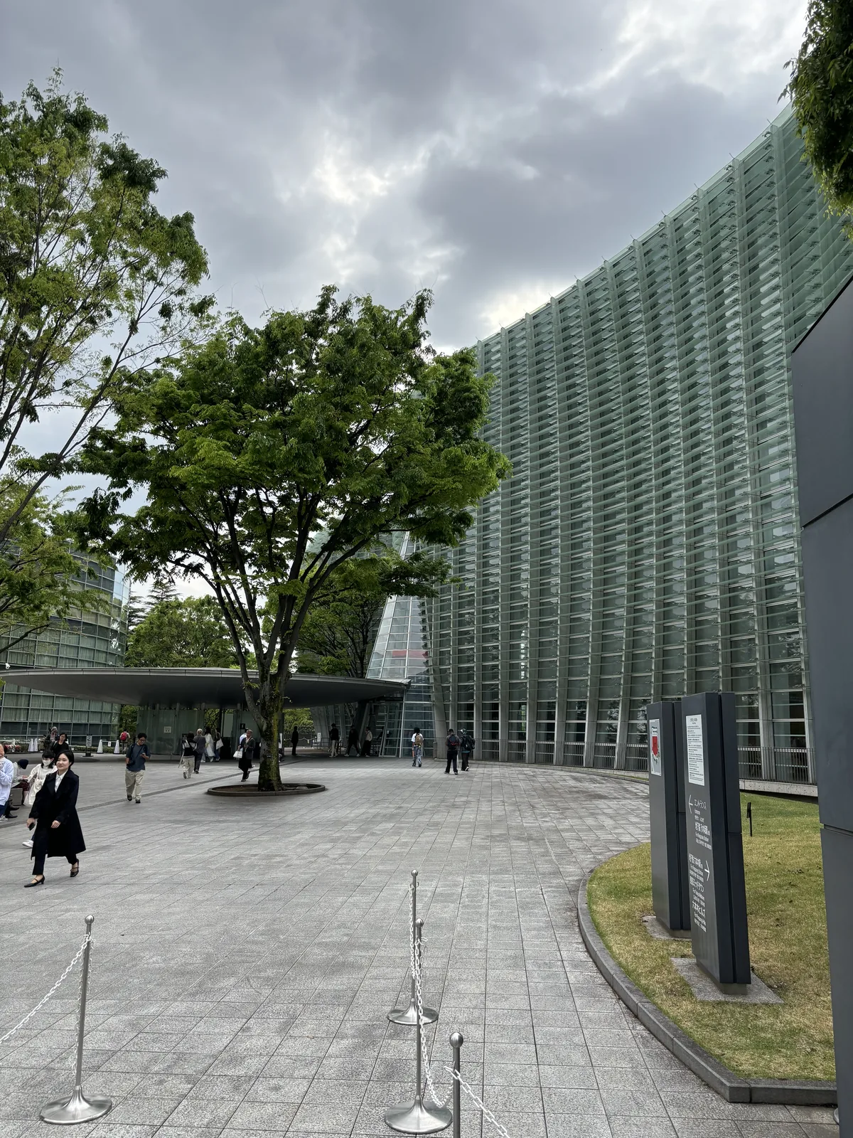 正門入口前的鋪石廣場，中央有一棵樹，右側是向外彎曲的玻璃 curtain wall，遠處可見入口前的雨庇與排隊的行人