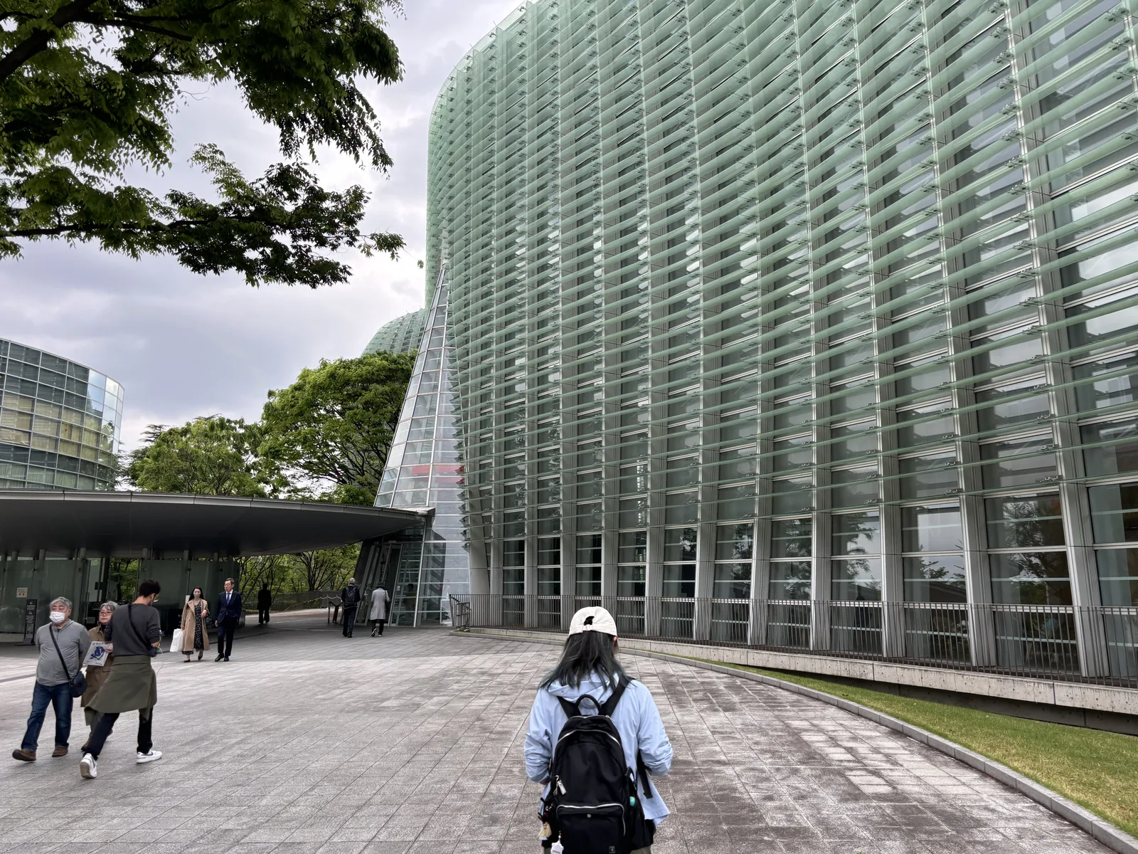 沿著玻璃 curtain wall 的鋪石步道，步道右側是草坪，玻璃面向前方延伸並在遠處與一座玻璃圓錐相接，圓錐前方是一片平坦的雨庇