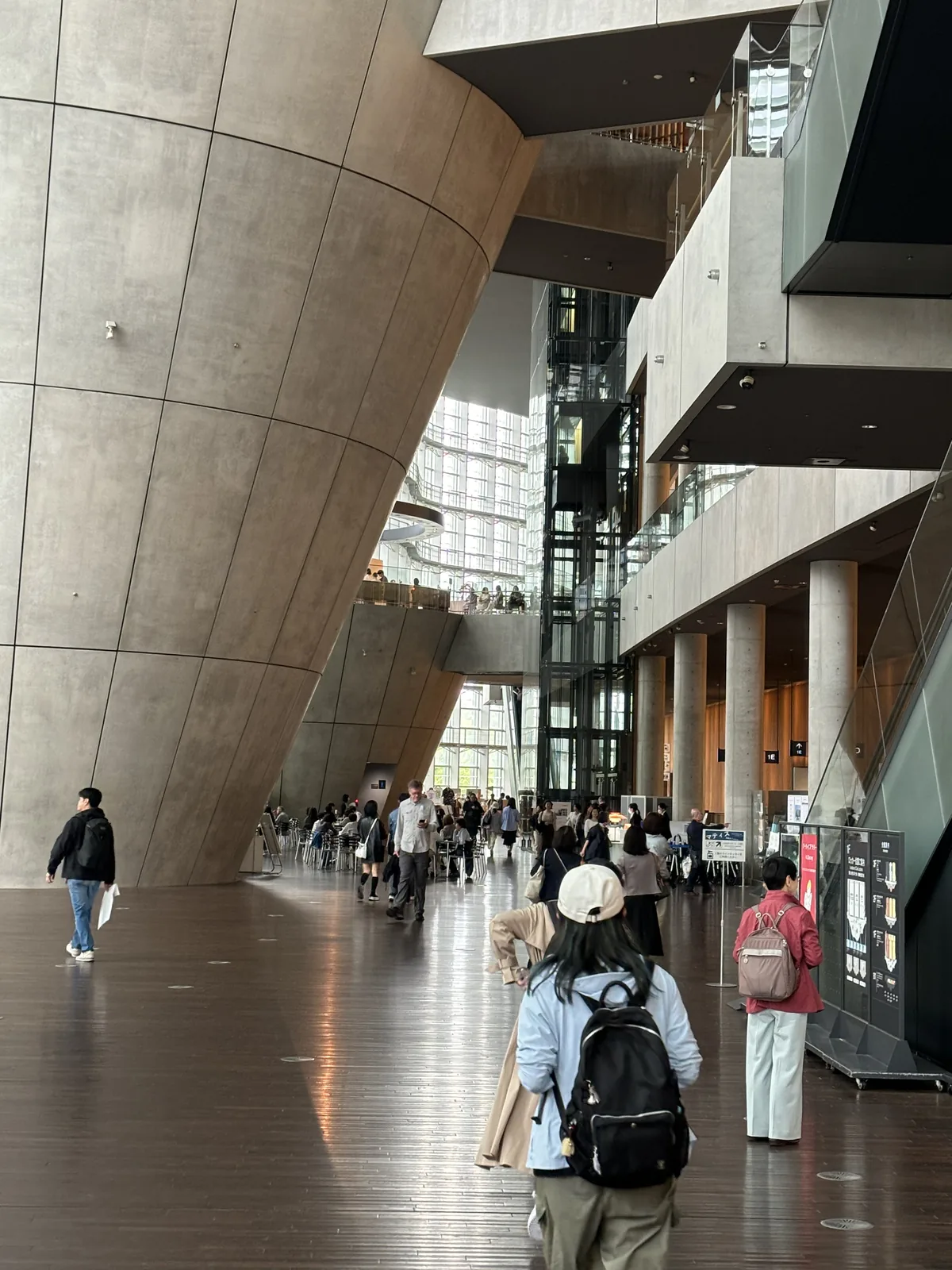 大廳中央，左側是大型 cone 的清水混凝土曲面，中央是玻璃電梯井與上層的走道，右側排列著清水混凝土圓柱，地板上有參觀者走動