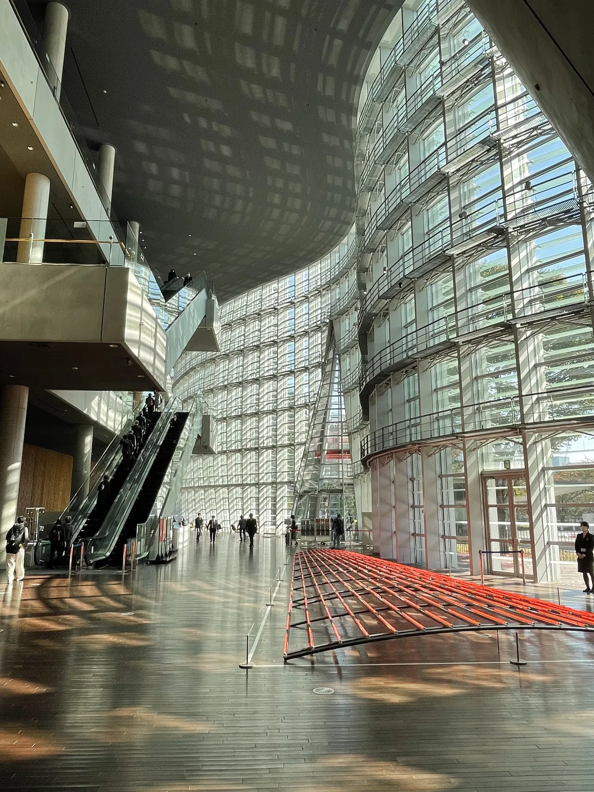 大廳的東西向長軸，右側是玻璃 curtain wall，louver 的影子以格子狀落在木地板上，中央地面放置著一件紅色的線狀裝置，遠處是手扶梯與入口