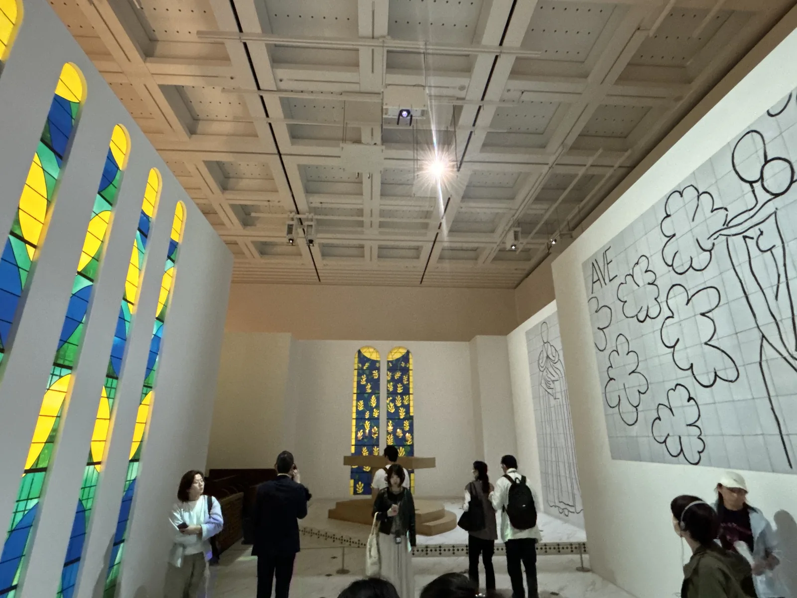 展示室內重現的旺斯禮拜堂內部，左側是排列著黃、綠、藍色彩繪玻璃窗的白牆，正面是祭壇與兩扇彩繪玻璃窗，右側牆面是黑白線描的磁磚壁畫，上方是展示室的格子天花