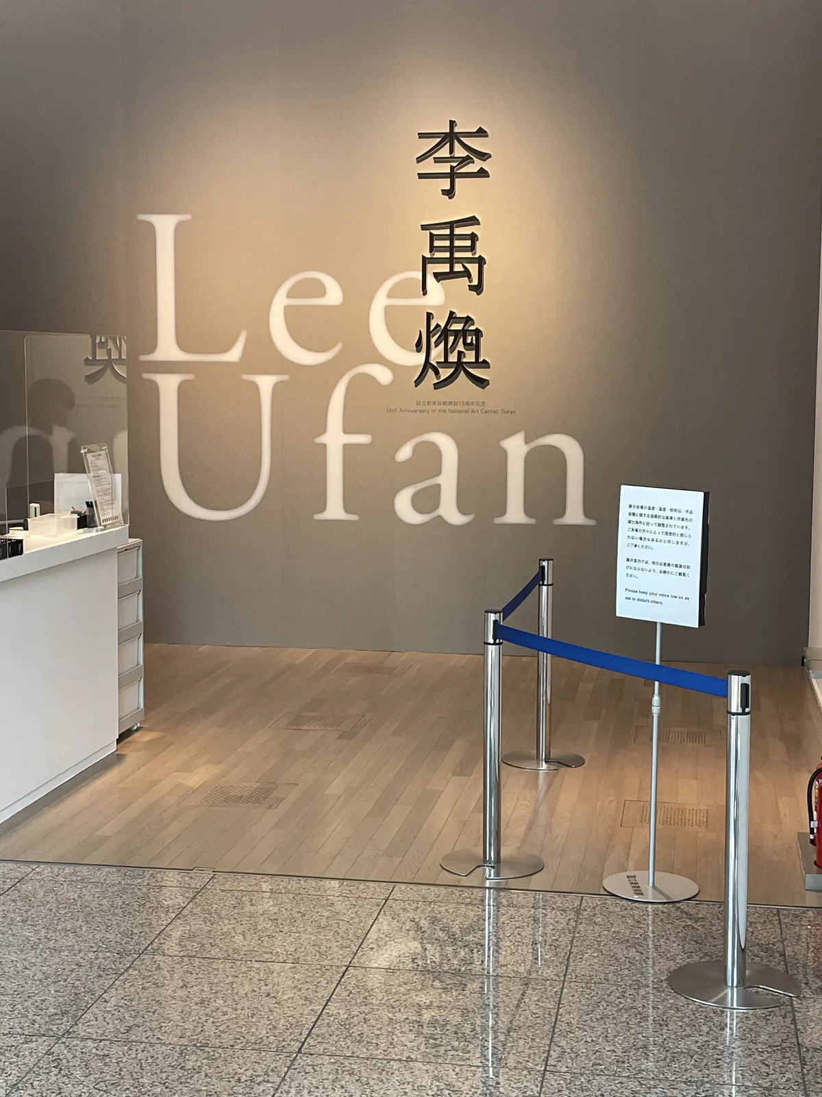 企劃展示室 1E 的入口，灰色牆面上以大字寫著李禹煥的中文名與英文名，左側是白色的接待台，前方以藍色伸縮帶設置排隊動線，地板是淺色石材