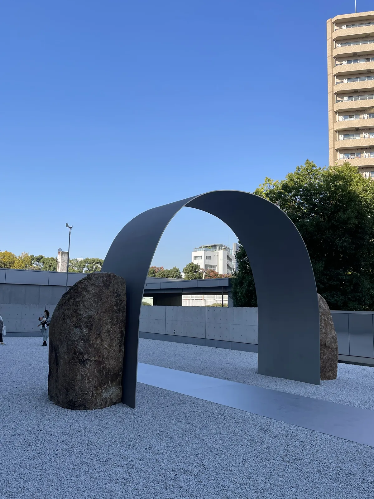 野外展示場，白色碎石鋪面上立著一道彎曲成拱形的鋼板，兩側各放置一塊自然石，背景是清水混凝土的擋土牆、樹木與住宅大樓