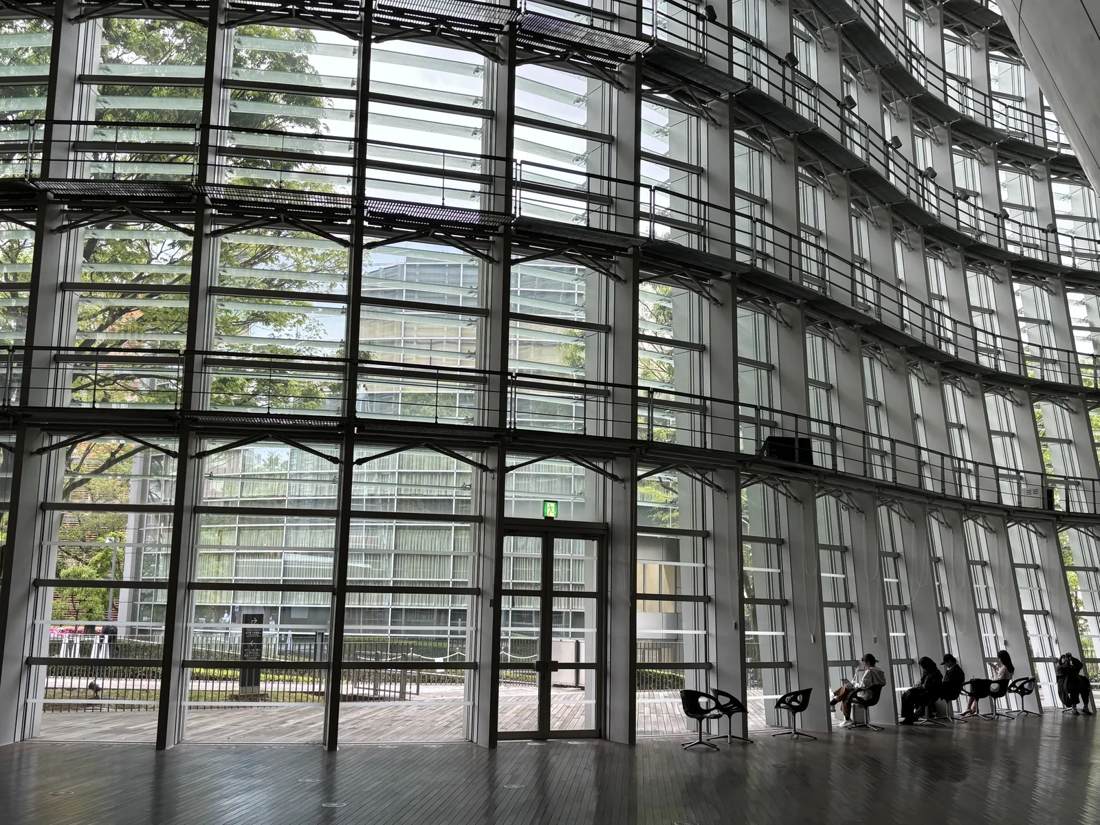 自館內望向玻璃 curtain wall，垂直的玻璃面外側排列著多層水平的玻璃 louver，鋼構的豎框與橫梁形成格子，下方有一扇通往外部木甲板的門，窗邊放著幾張椅子