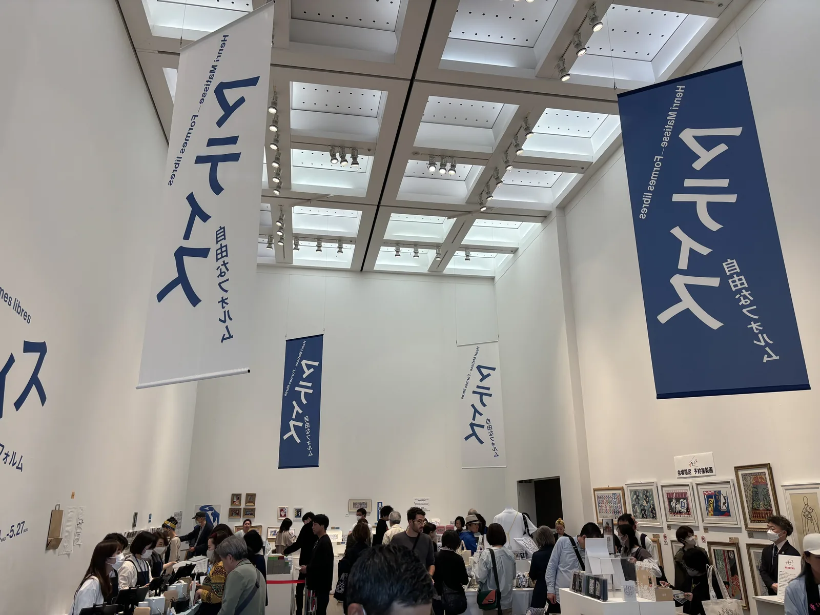 展示室內設置的展覽特設商店，挑高的白色空間上方懸掛著藍白色的展覽布條，天花是格子狀的方格，下方排列著商品台與排隊結帳的人群