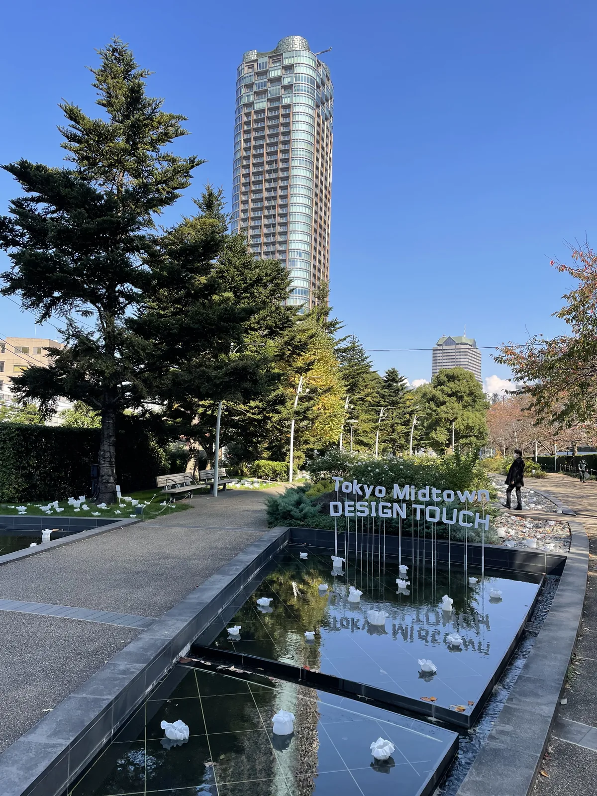 東京中城庭園的步道，左側是長方形的水池，池中與池畔放置著白色的裝置作品，水池旁立著英文標誌，背景是松樹與一棟高層住宅大樓