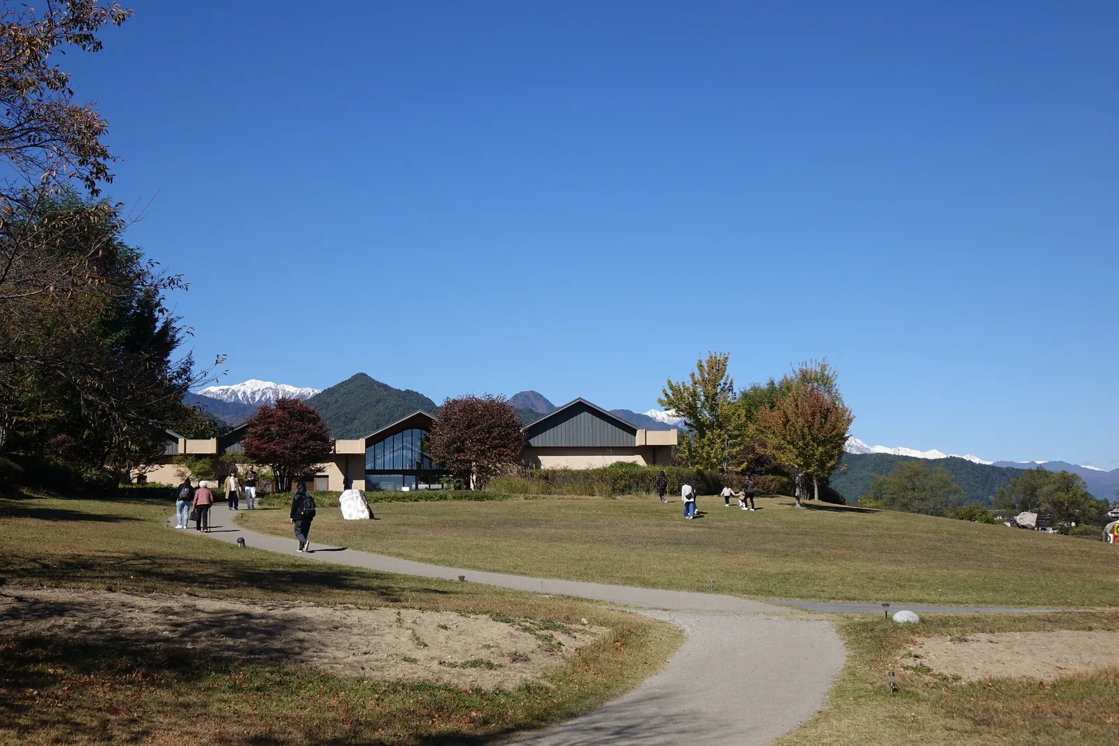 公園的步道蜿蜒穿過草地，遠方是一列低矮的山牆建築與覆雪的山峰，步道上有數位行人