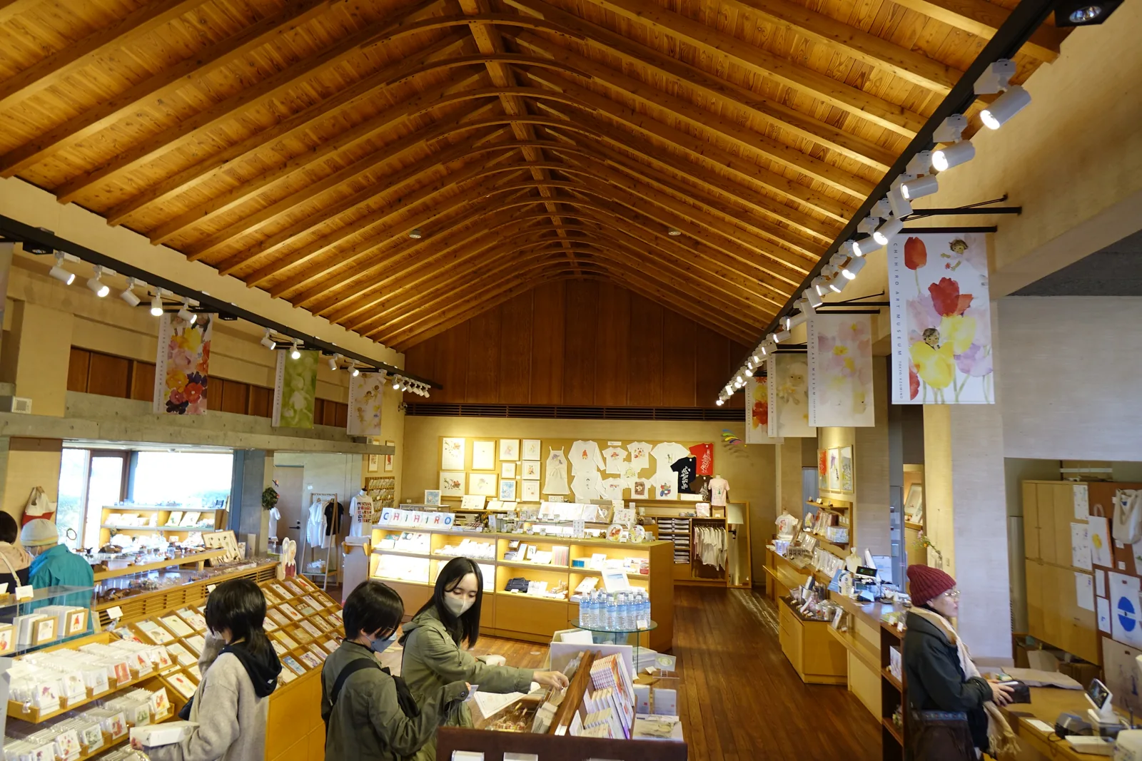 商店內部，山牆形的木製屋架之下是木製的陳列台與展示架，數位來館者正在選購，牆上掛著大幅的畫作