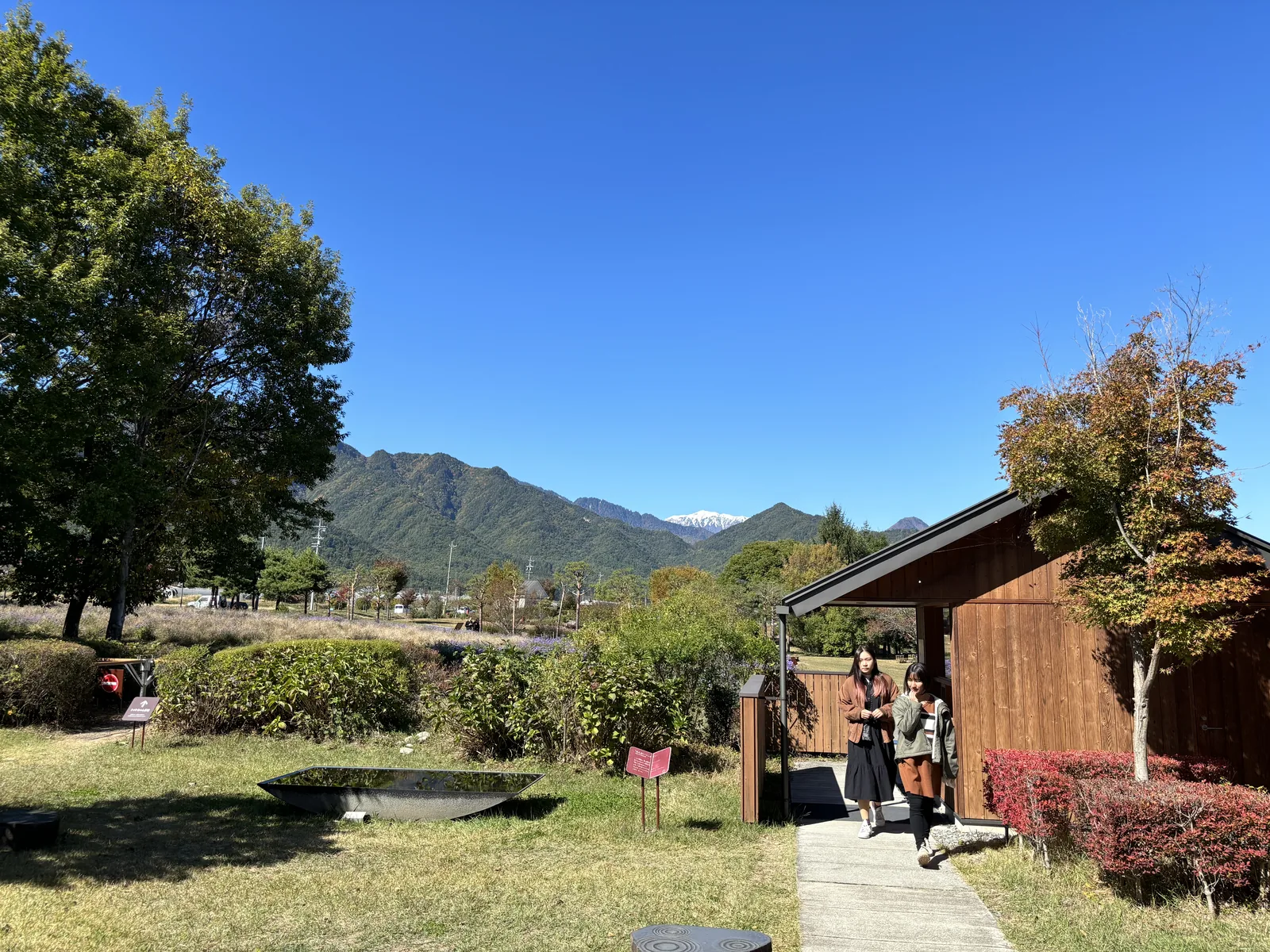 公園一角的木造小屋，前方是草地與花圃，背景是遠方的群山與藍天，小屋前有兩位來館者