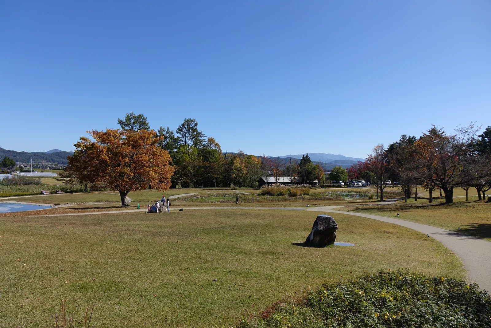 公園的大片草地，中央有一塊黑色的石頭，周圍是轉紅的樹木與遠處的低山，天空晴朗