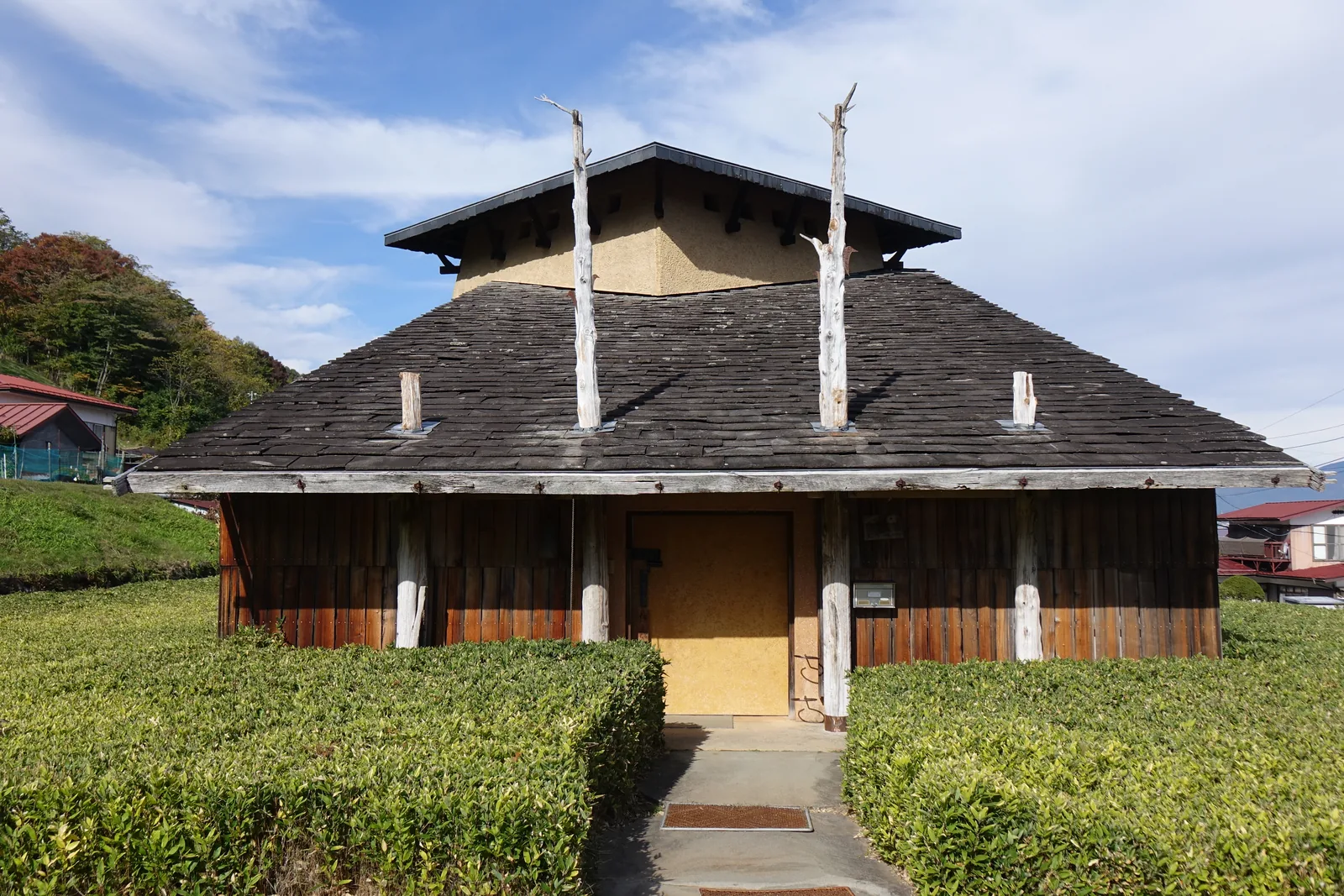 坡地上的單層建築，深灰色石板斜屋面，兩根灰白色樹幹自雨庇穿出並高過後方二層量體的屋頂，立面為直拼木板，入口門為土黃色，前方是修剪整齊的綠籬與石階