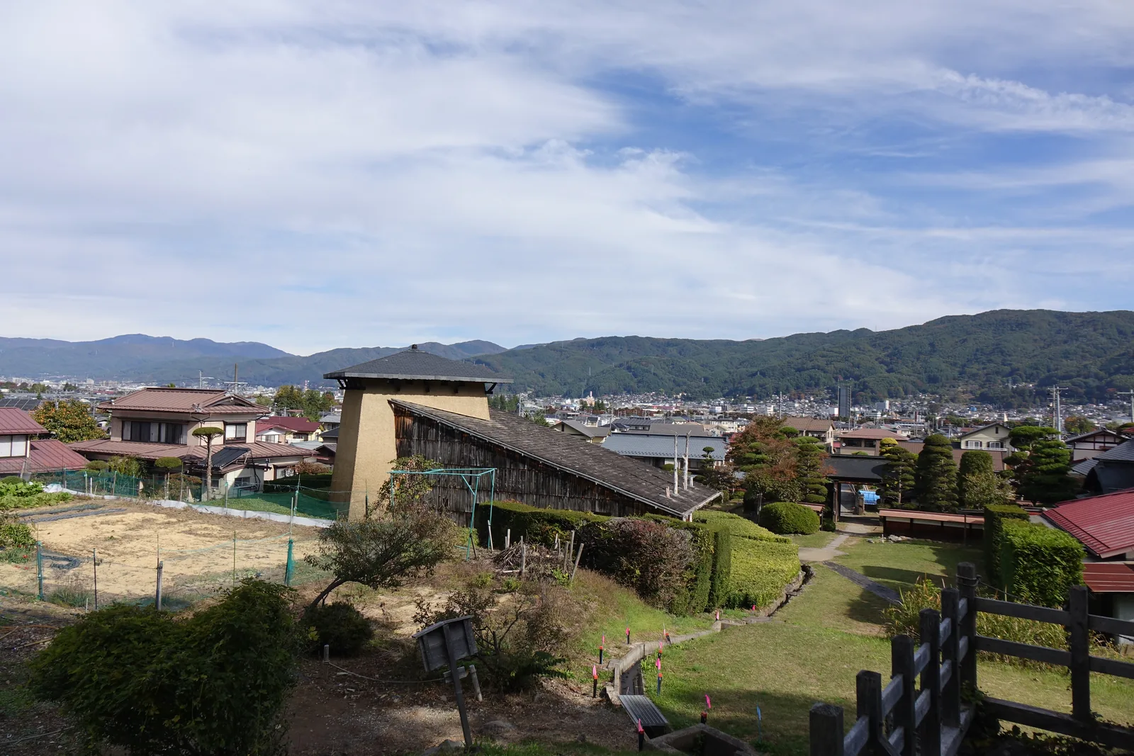 更廣的俯瞰視角，史料館位於畫面中央偏左，周圍是住宅的屋頂、庭樹與菜園，遠方是諏訪盆地的市街與山並