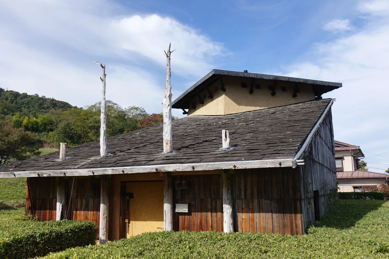 自斜前方看史料館，石板斜屋面向右下降，兩根高柱與兩根短柱穿出屋面，後方是土黃色的二層量體與深色方形屋頂，側面山牆為直拼木板
