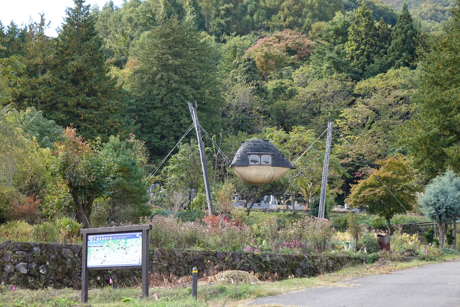 坡地的道路旁，兩組高柱之間以鋼索懸吊著一個船形的小屋，下方是花圃與石砌擋土牆，路邊立有步道的案內板，後方是樹林
