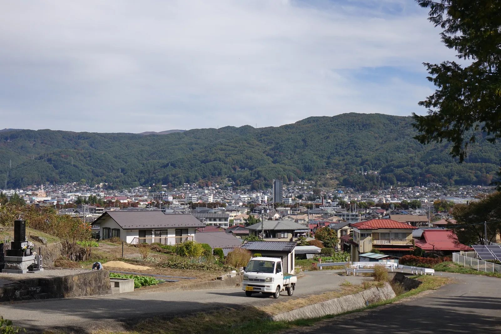 坡地上的道路與聚落，前景是農地與一輛輕型貨車，中景是住宅的屋頂，遠方是盆地的市街與連綿的山