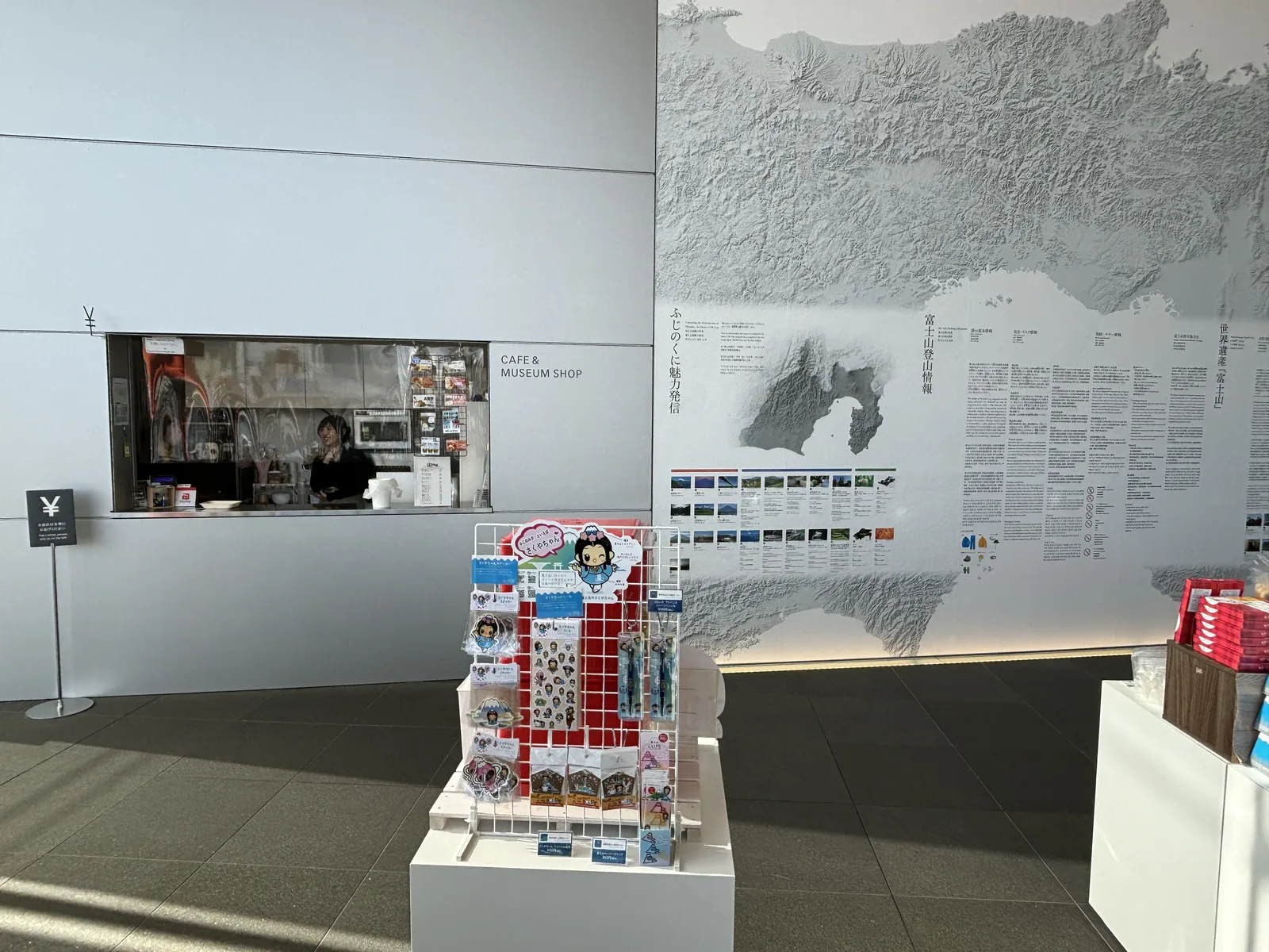 白色牆面上開著一個橫長的櫃檯窗口，上方標示 CAFE & MUSEUM SHOP，右側牆面是靜岡縣的地形浮雕圖與說明文字，前方是商品陳列台