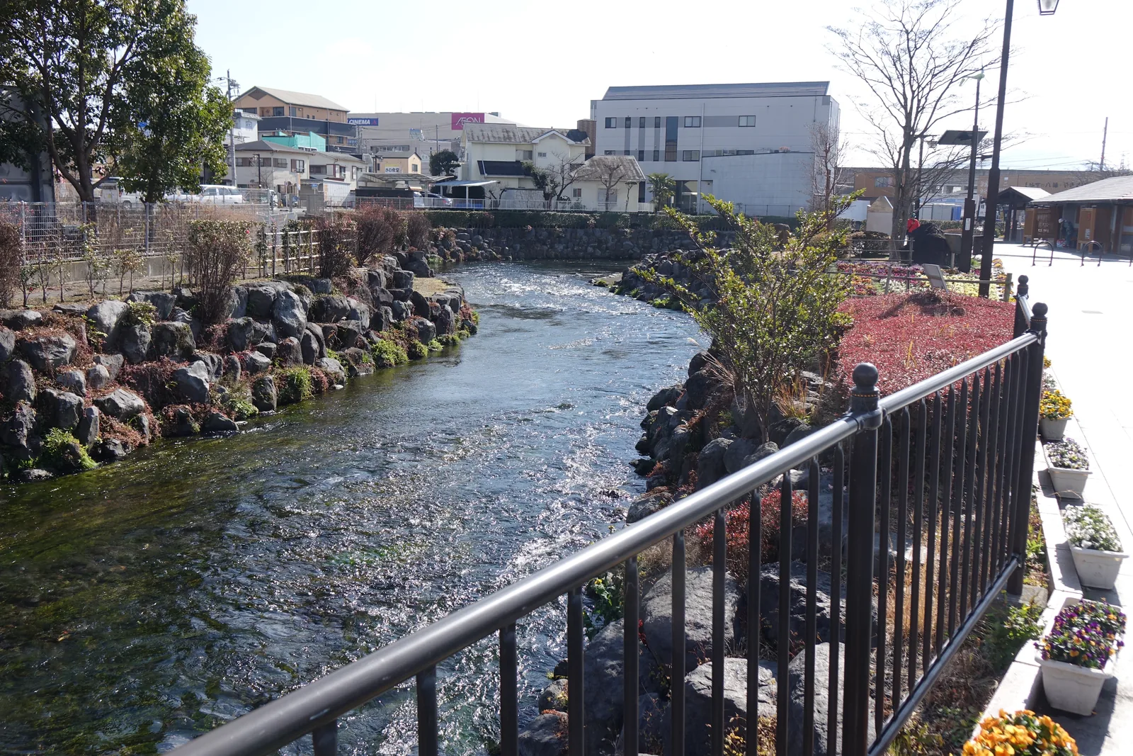 流經市區的小河，水流湍急，兩岸以黑色的熔岩石砌成護岸，右側是紅葉的矮灌木與花圃，遠方是市街建築