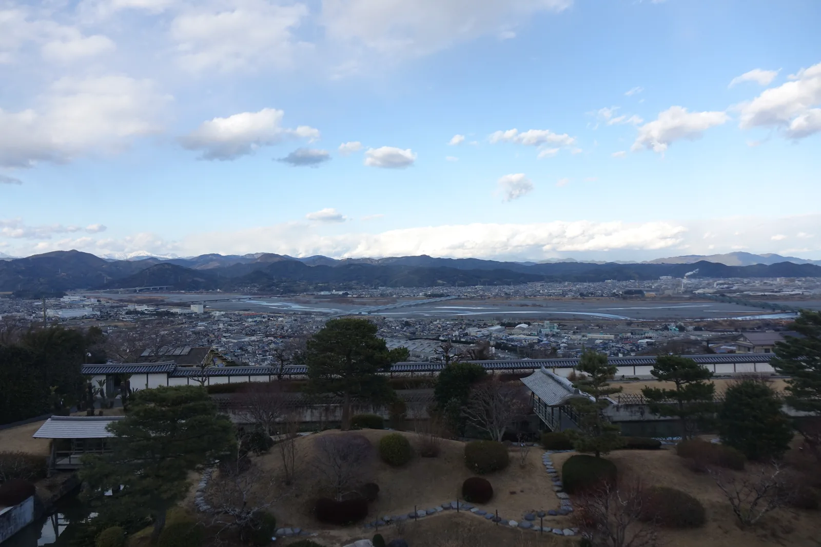 自高處眺望，前景是有水池、築山與松樹的庭園，中景是住宅與茶田，遠方是寬闊的河灘、市街與連綿的山