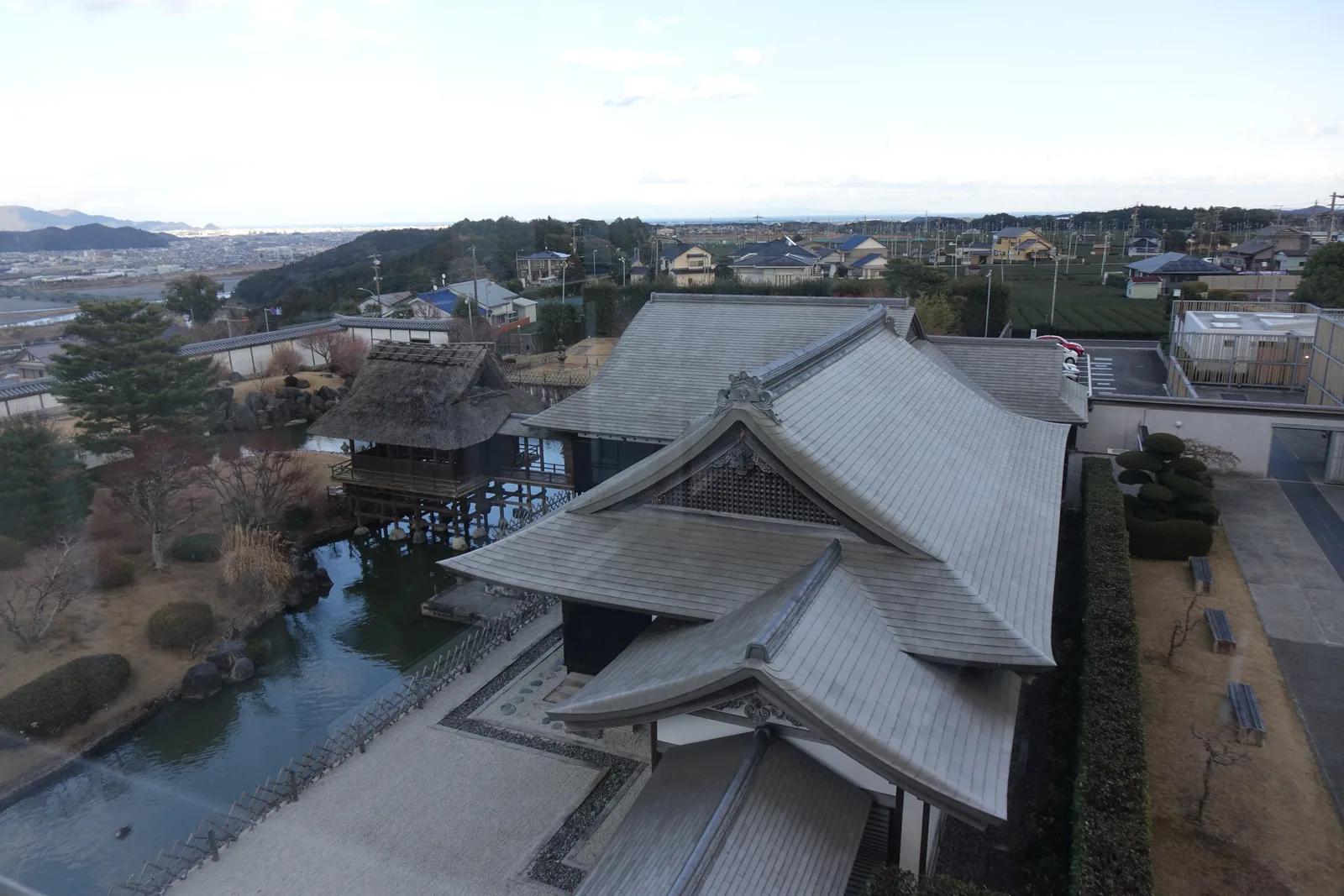 自高處俯瞰，中央是灰色屋頂重疊的茶室建築群，左側是水池與架在池上的茅草頂小亭，後方是住宅、茶田與遠方的海岸線