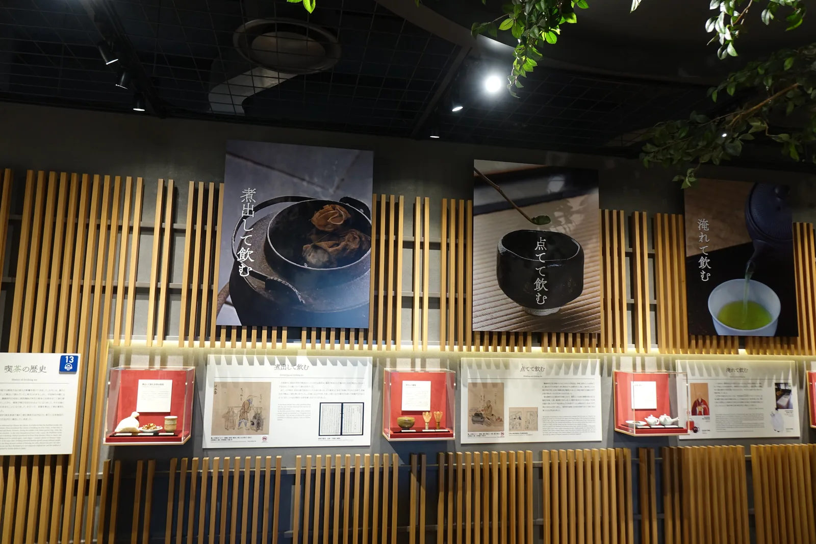 展示牆以成組的淺色直立木條構成，木條前掛著三幅茶的照片與紅色的展示盒，上方的黑色天花垂下茶樹的枝葉