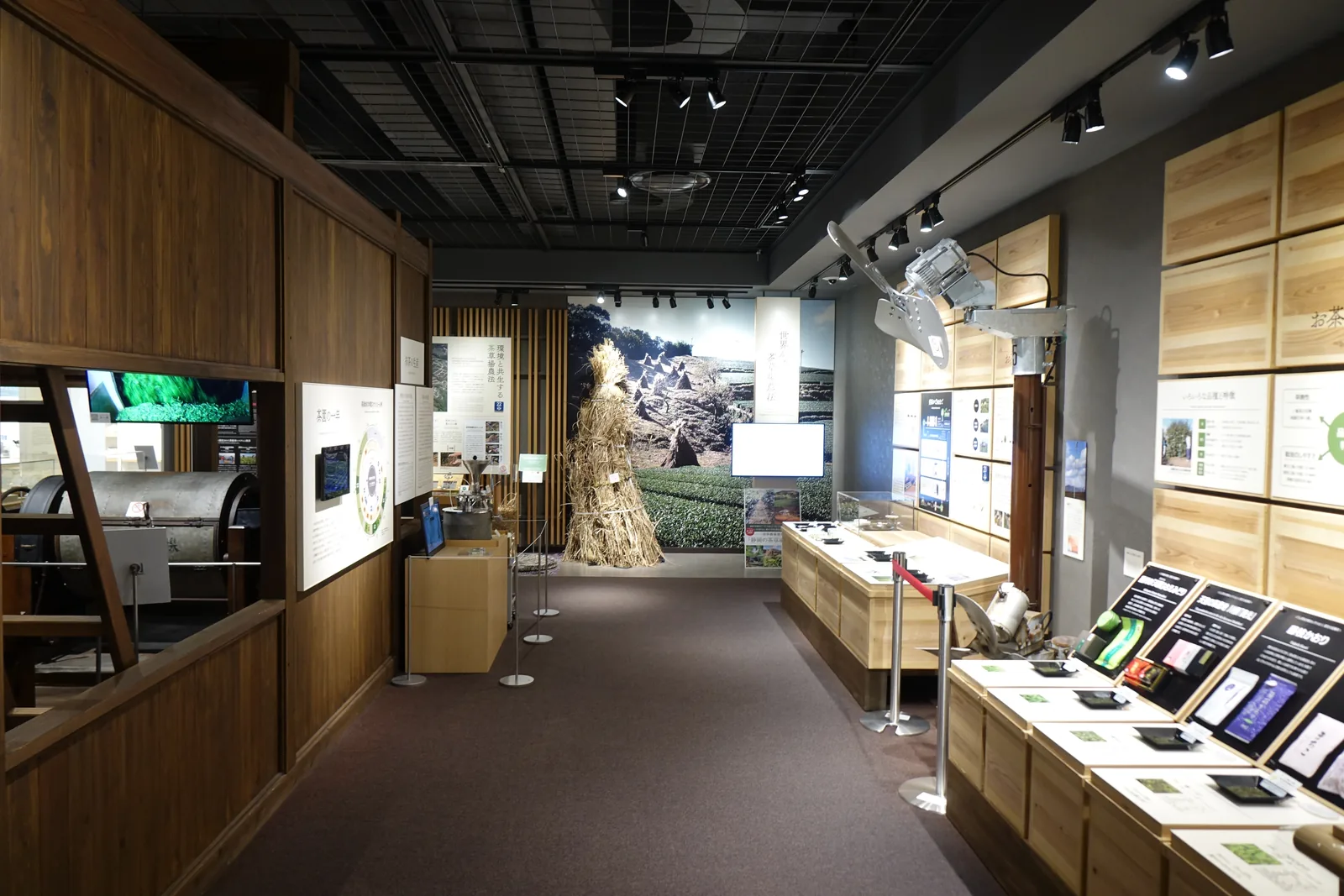 二樓的展示通道，左側是深褐色的木板牆，右側是淺色木格的圖版牆與展示台，盡端立著一束乾草與茶田的大幅照片