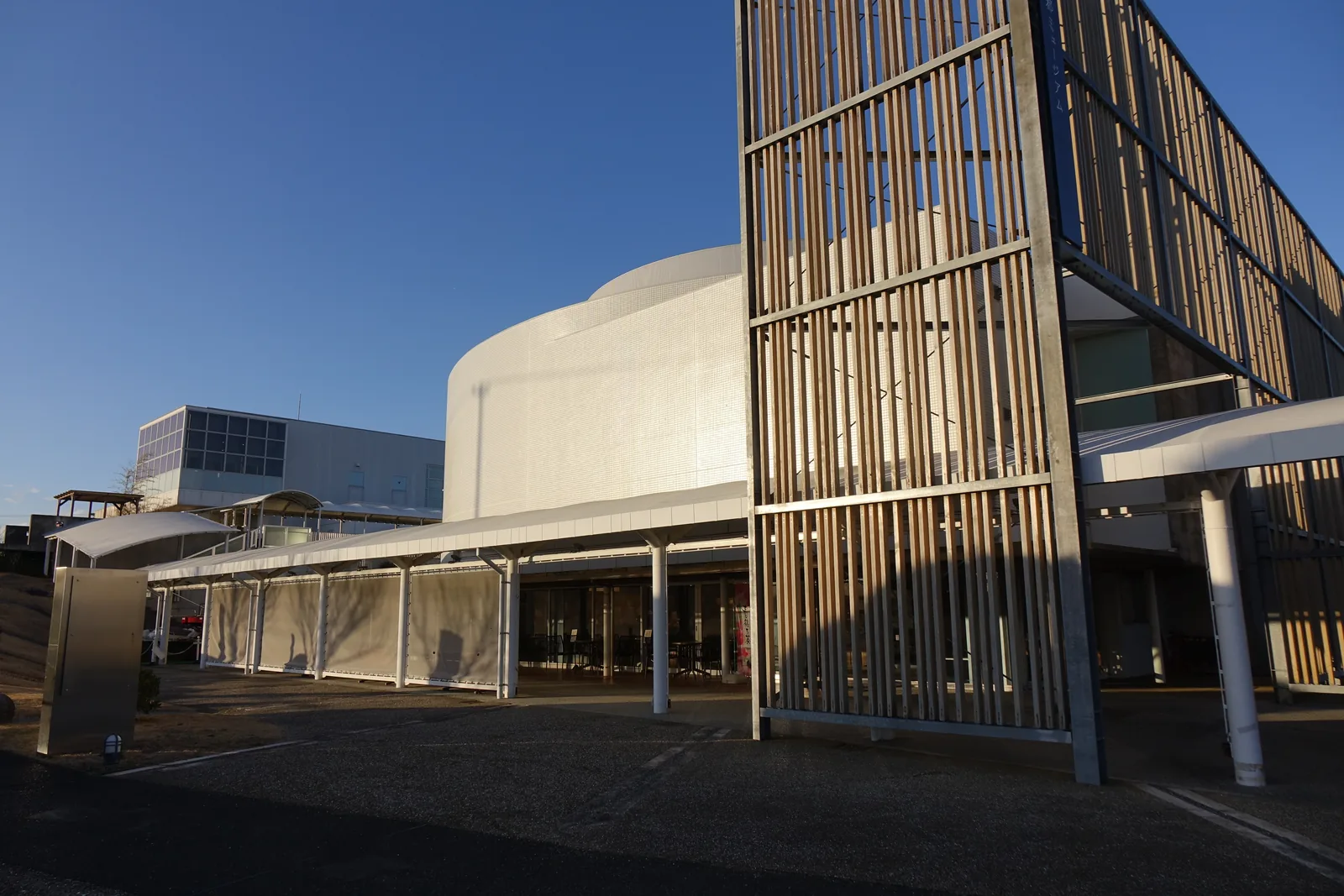 近看建築的轉角，直立的木格柵固定在鍍鋅鋼構架上，高出後方的白色雨庇，格柵後是白色的圓筒形量體，天空深藍