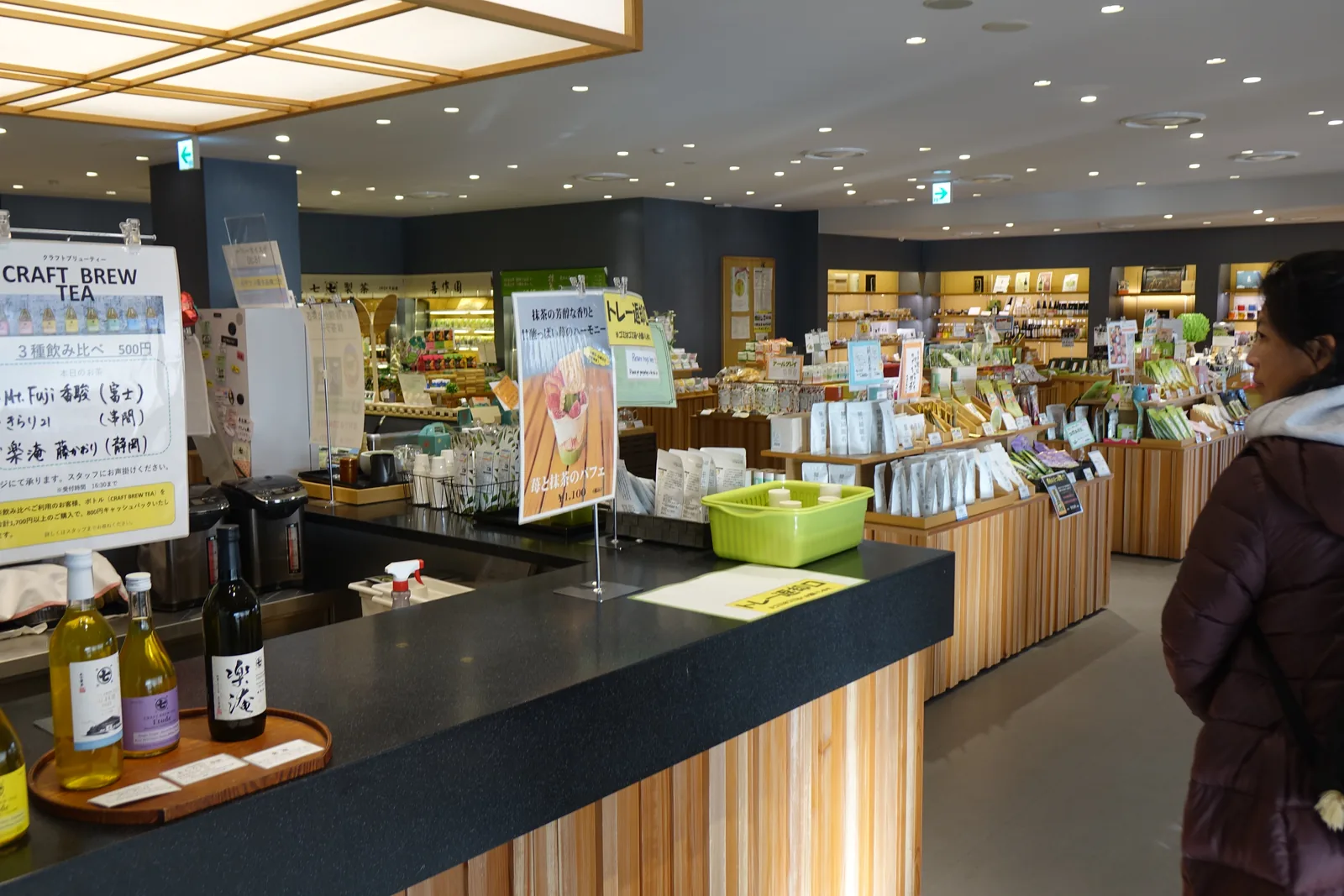 商店內部，上方是木格子的大型方形燈箱，前方是黑色台面的櫃檯與成排的茶飲瓶，後方是陳列茶葉商品的低矮木台，一位訪客走過