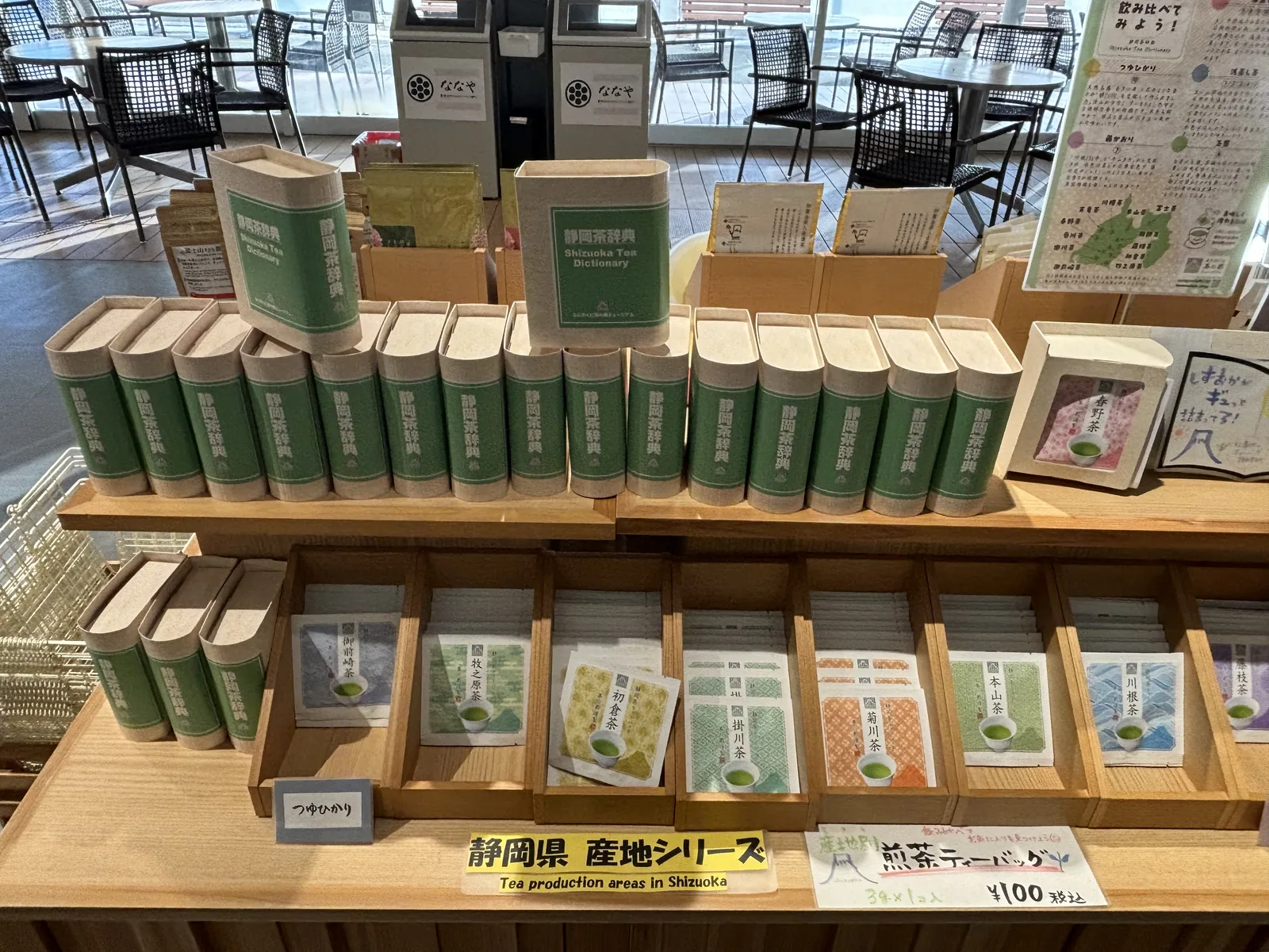 商店的陳列台上擺著成排的綠色茶筒與茶包，立著靜岡縣產地系列的標示與手繪的產地地圖，後方的玻璃外是露台的桌椅