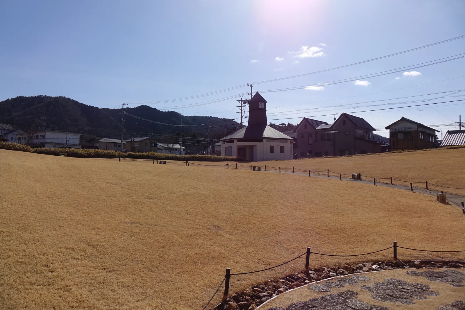 枯黃的草坡向下傾斜，坡底是圓形的石板鋪面，遠方是低層的住宅、一座有尖頂鐘塔的小屋與山丘