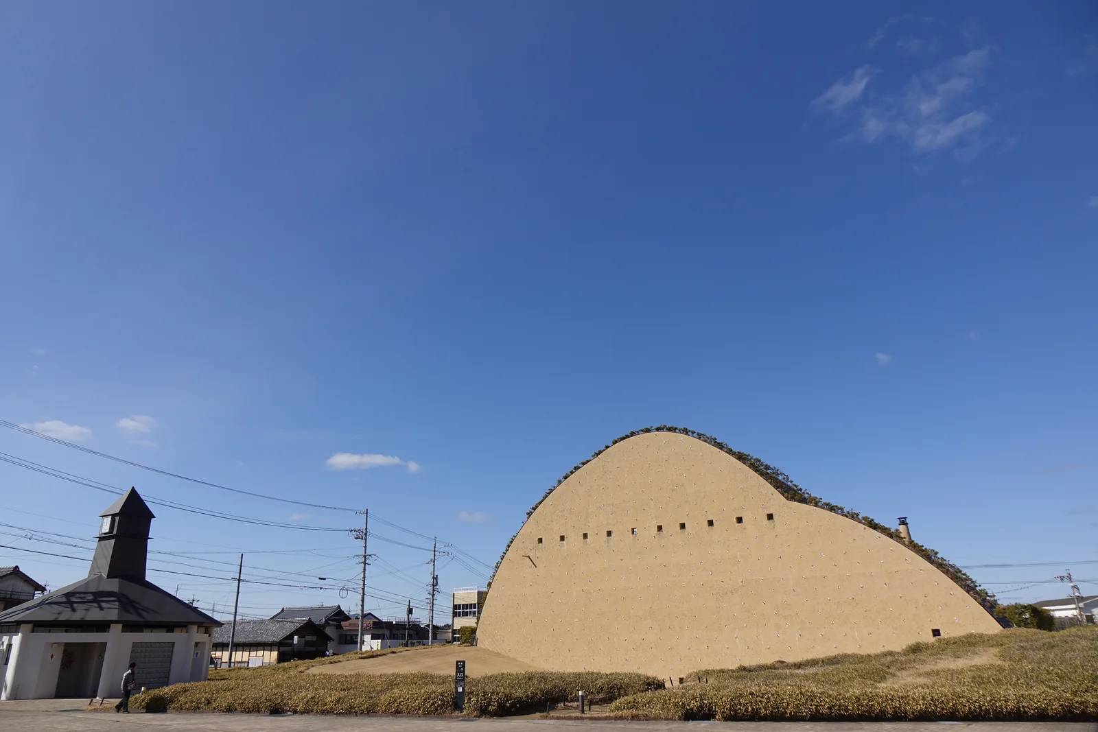 前景是一座白牆、深色屋頂、頂部設有鐘塔的小屋，後方是博物館土黃色的曲線牆面，天空深藍
