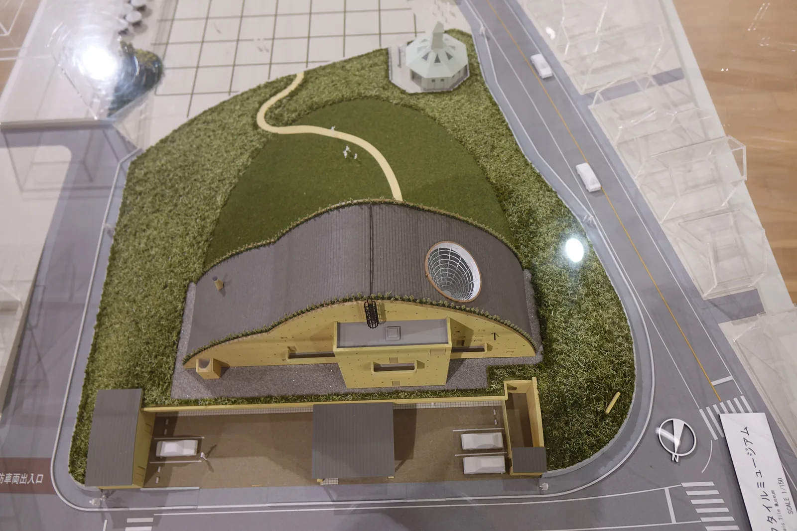 玻璃罩內的建築模型，中央是筒形拱屋頂的博物館，南側是研缽狀的草坡與弧形步道，角落有一座小屋，四周是道路