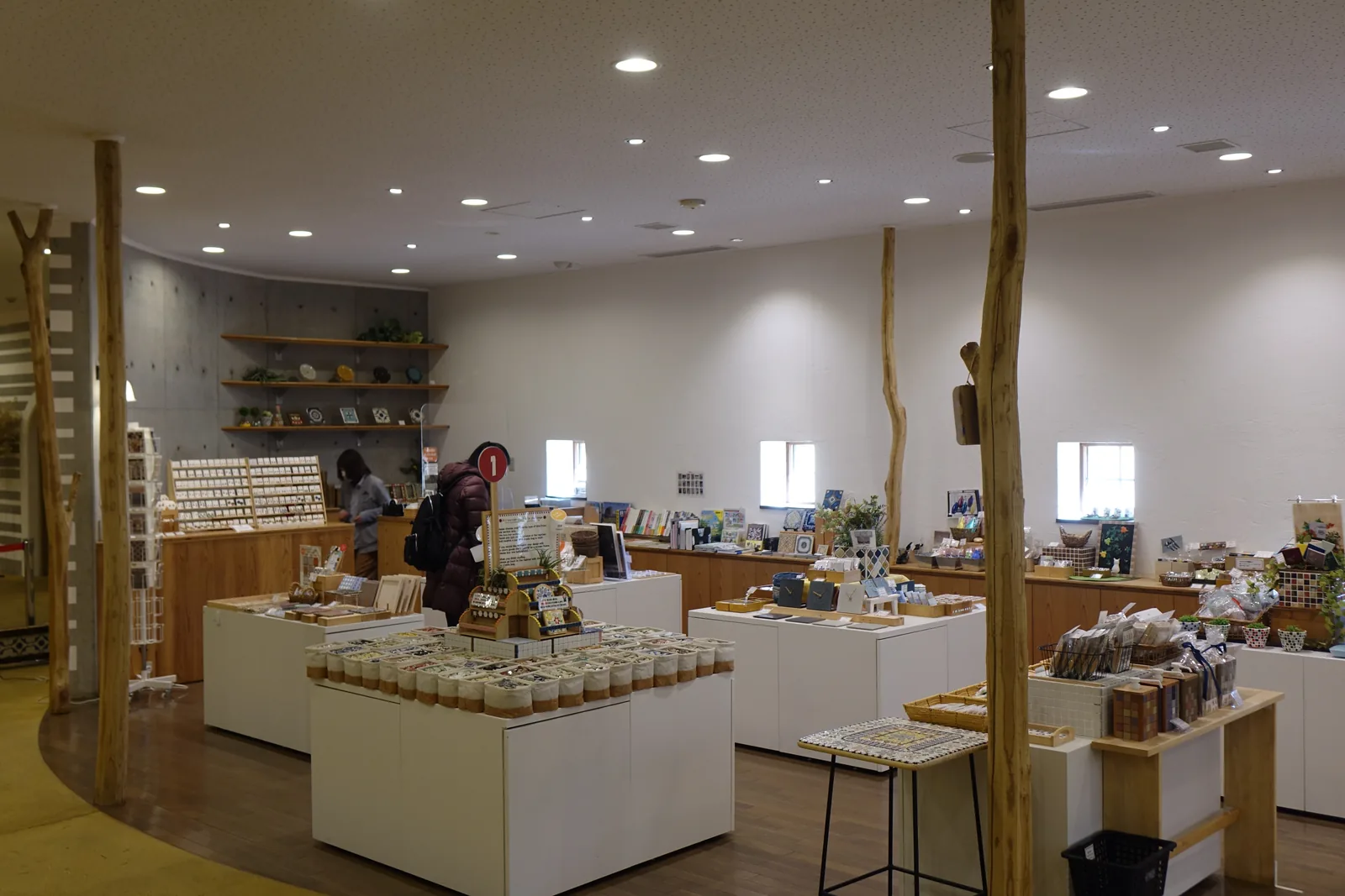一樓的商店，數根保留自然形狀的細木柱立在賣場內，白色的陳列台上擺滿瓶裝的磁磚與小物，後方是白牆、小方窗與清水混凝土牆上的木層架