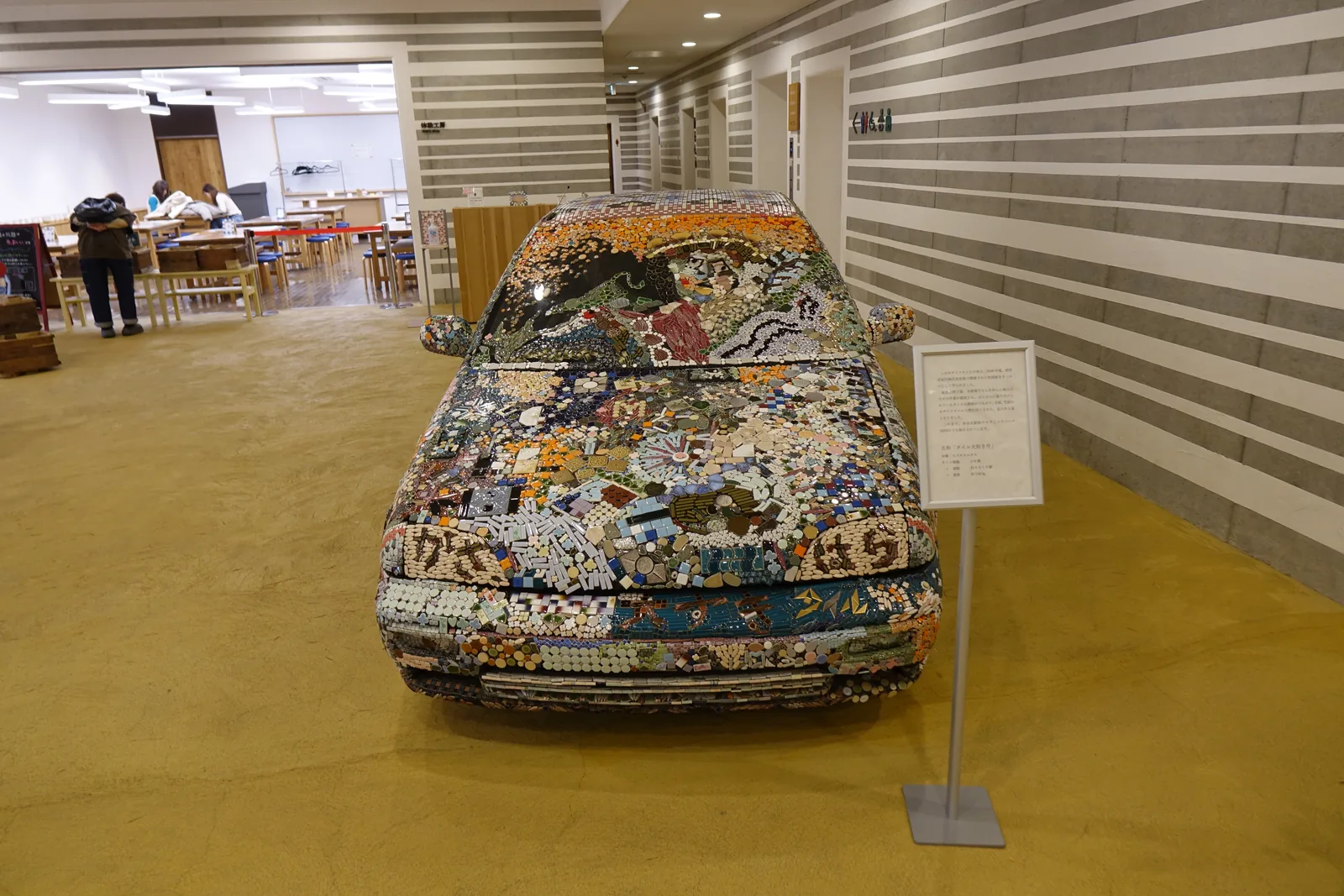 一樓大廳中展示的汽車，車身表面貼滿彩色的磁磚與陶片，後方是條紋牆面與體驗工房的開口