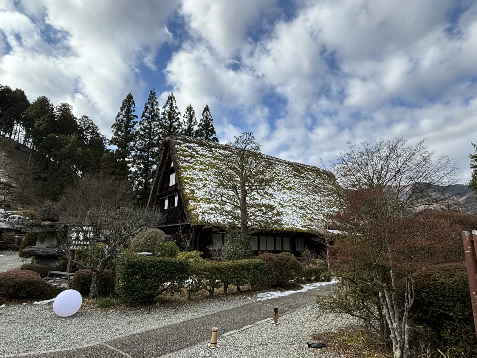 茅草屋頂的大型合掌造民家，屋頂積著薄雪並長有青苔，山牆面可見數層的窗，屋前是石燈籠、修剪過的灌木與一塊寫著村名的木牌，後方是杉林