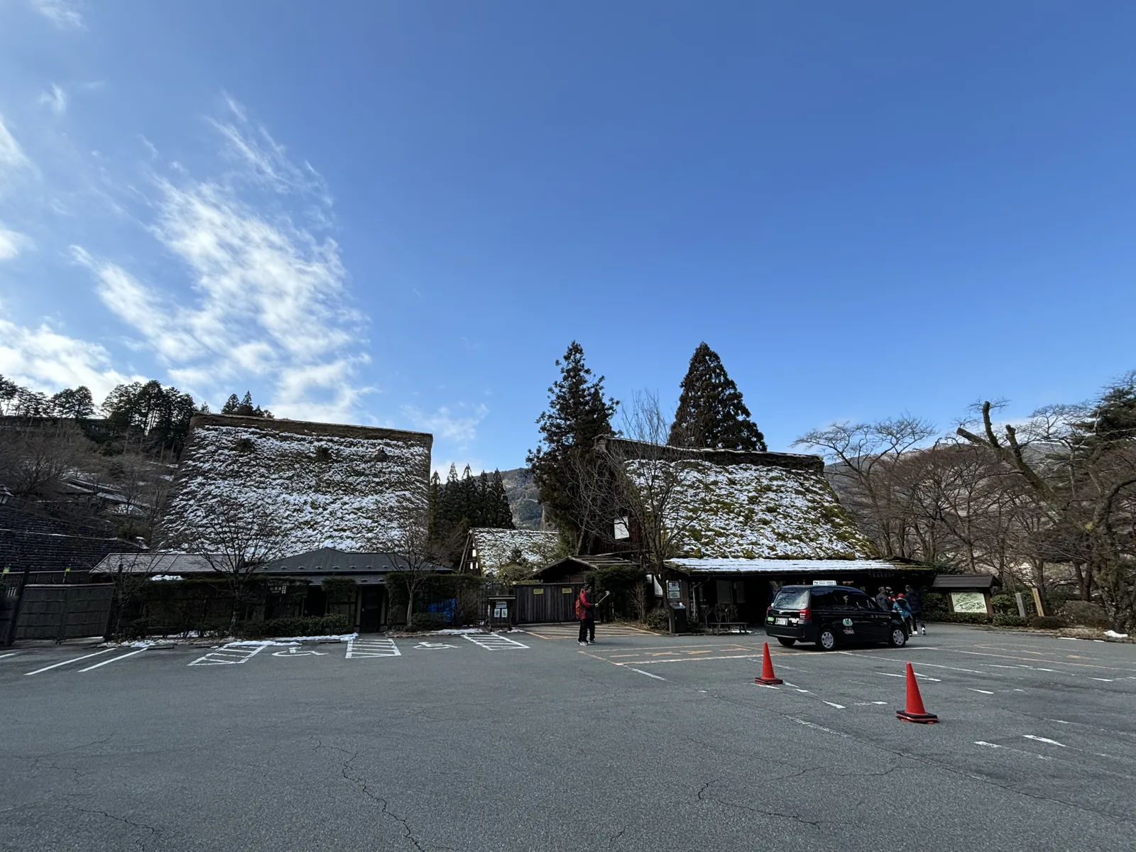 寬闊的停車場後方並排著兩棟茅草屋頂的建築，屋頂積著薄雪，其間與後方是數棵高大的杉樹，天空晴朗