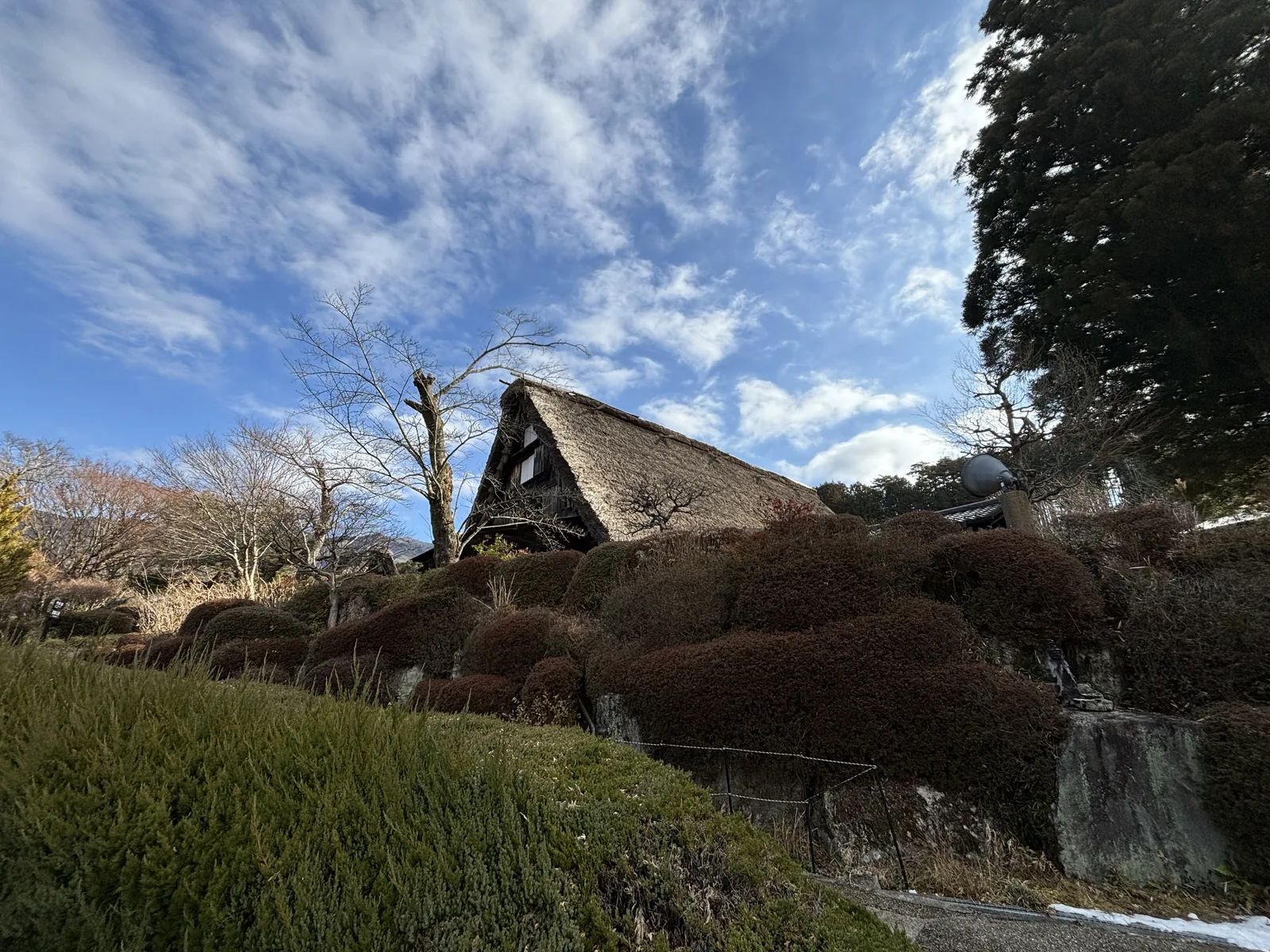 兩棟茅草屋頂的民家隔著灌木相對，左側一棟露出山牆的窗，右側一棟以長邊朝向前方，遠方是連綿的山