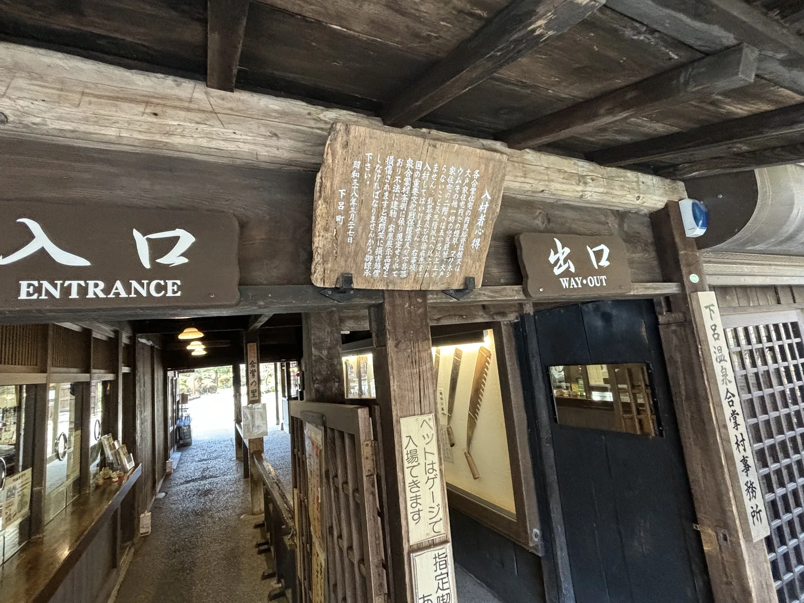 入口建築的內部通道，上方是深褐色的木樑與樓板，樑上掛著入口與出口的木牌與一塊寫滿文字的木板，通道盡端明亮，可見村內的道路