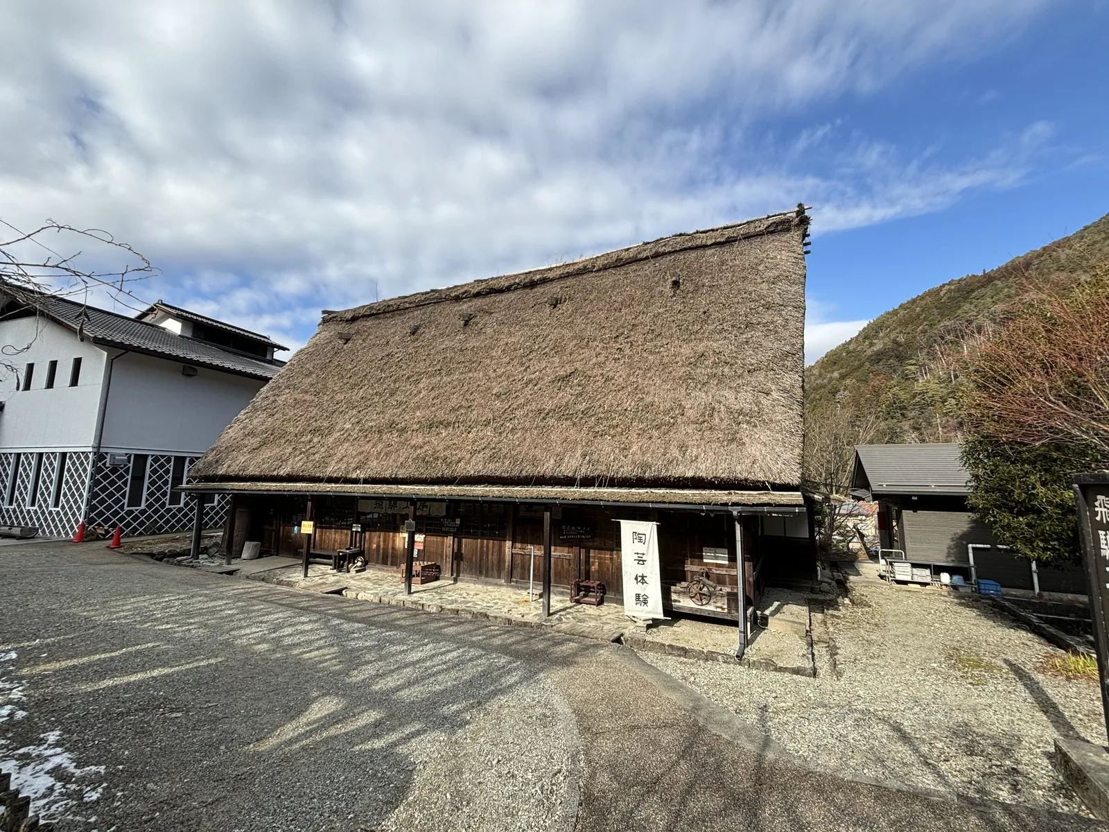 一棟長條形的茅草屋頂建築，屋頂的斜面很大，簷下立著寫有陶藝體驗的看板，左側是白牆黑瓦的建築，前方是碎石鋪面的廣場