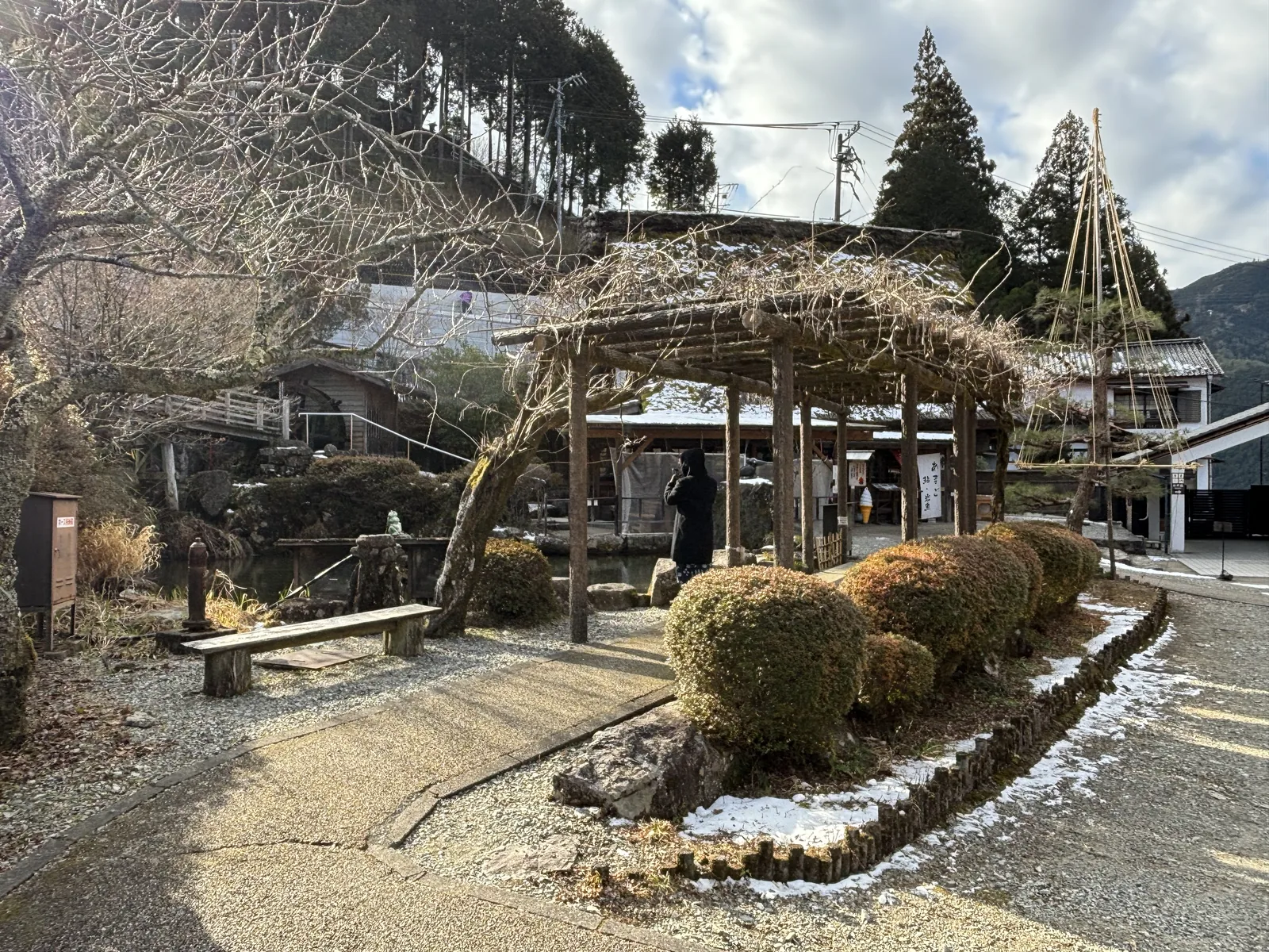碎石步道旁的木造藤架，冬季只剩纏繞的枯藤，藤架旁是修剪成圓形的灌木與石組，後方是白牆的建築與一座以竹與繩搭成的防雪吊架