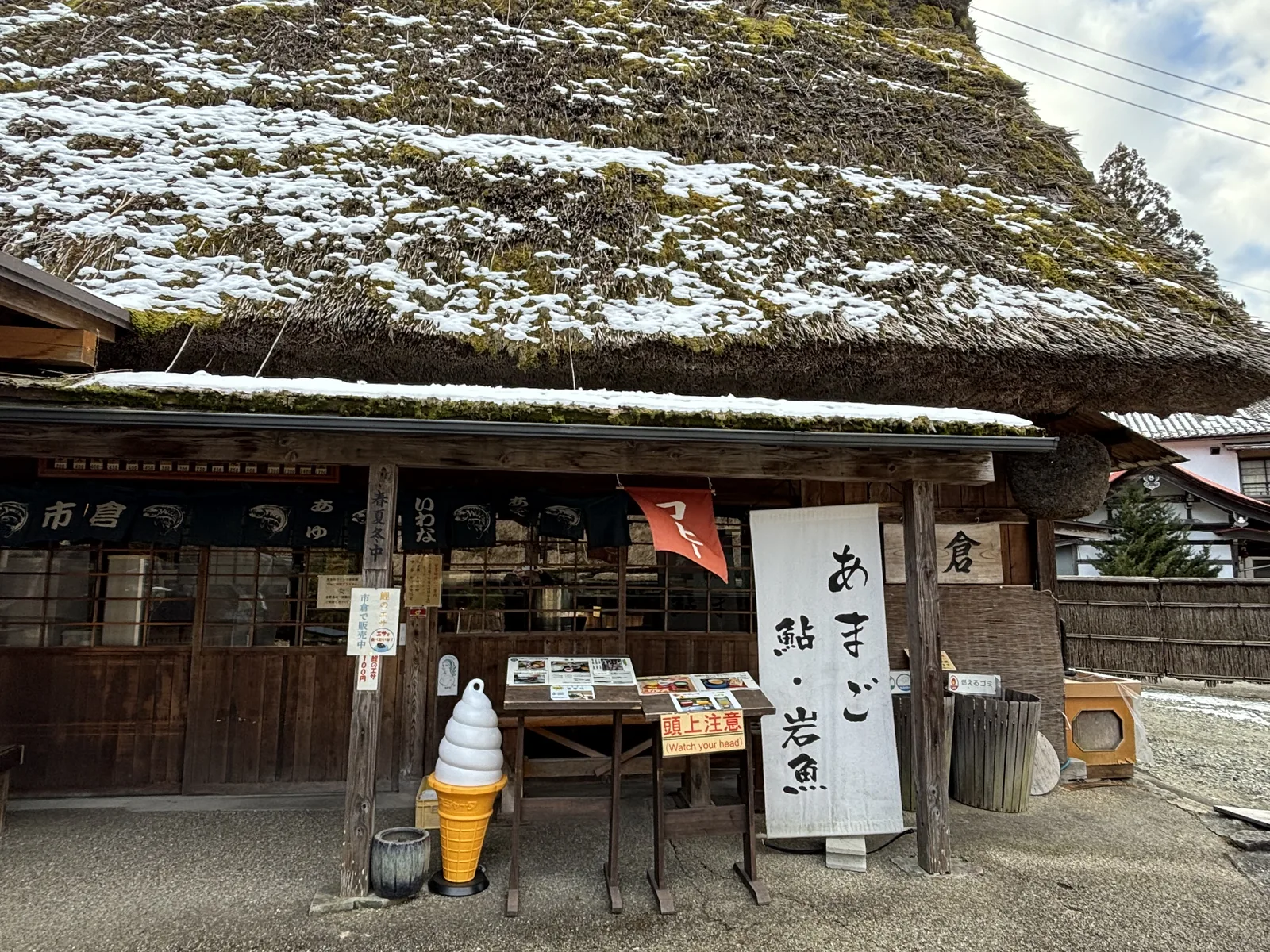 茅草屋頂建築的正面，屋頂積雪並長有青苔，簷下掛著印有店名的暖簾，門前立著手寫川魚名稱的白色立牌、菜單台與霜淇淋的模型