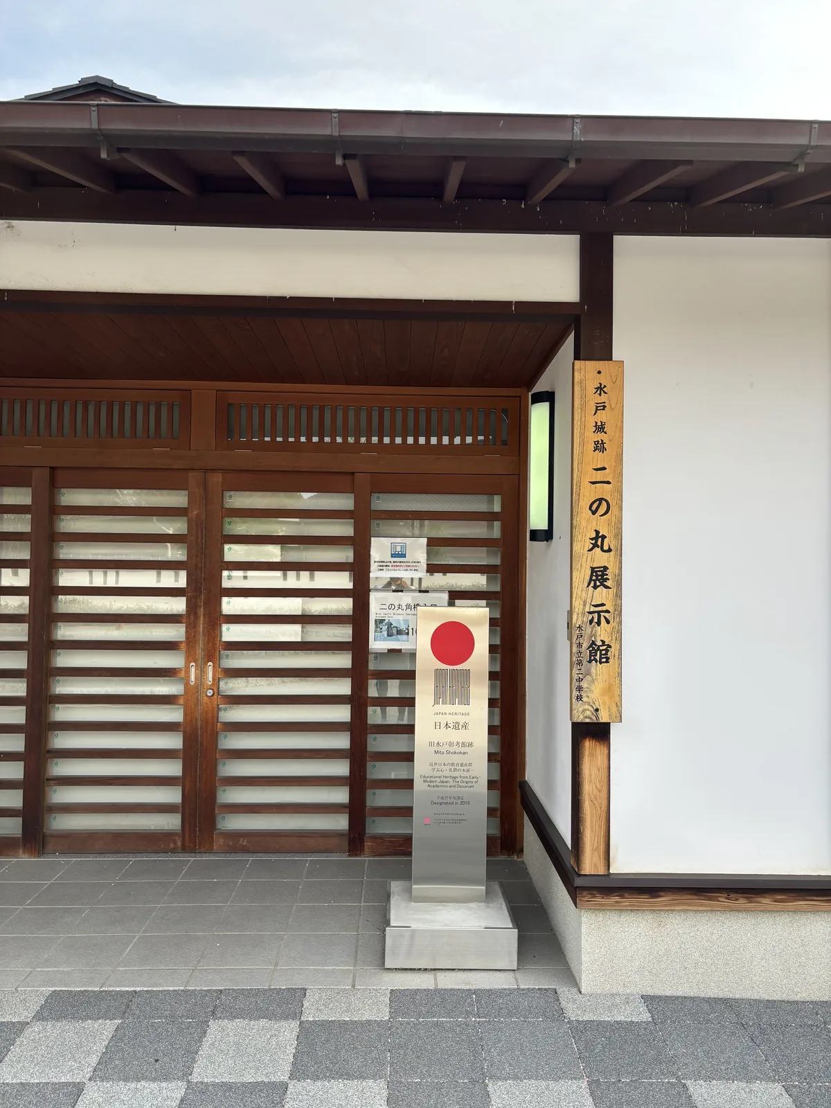 水戶城跡二之丸展示館的入口，深色木框的格子拉門上方有木製的出簷與露明的椽木，右側白牆上掛著直書的館名板，門前立著不鏽鋼的日本遺產標柱