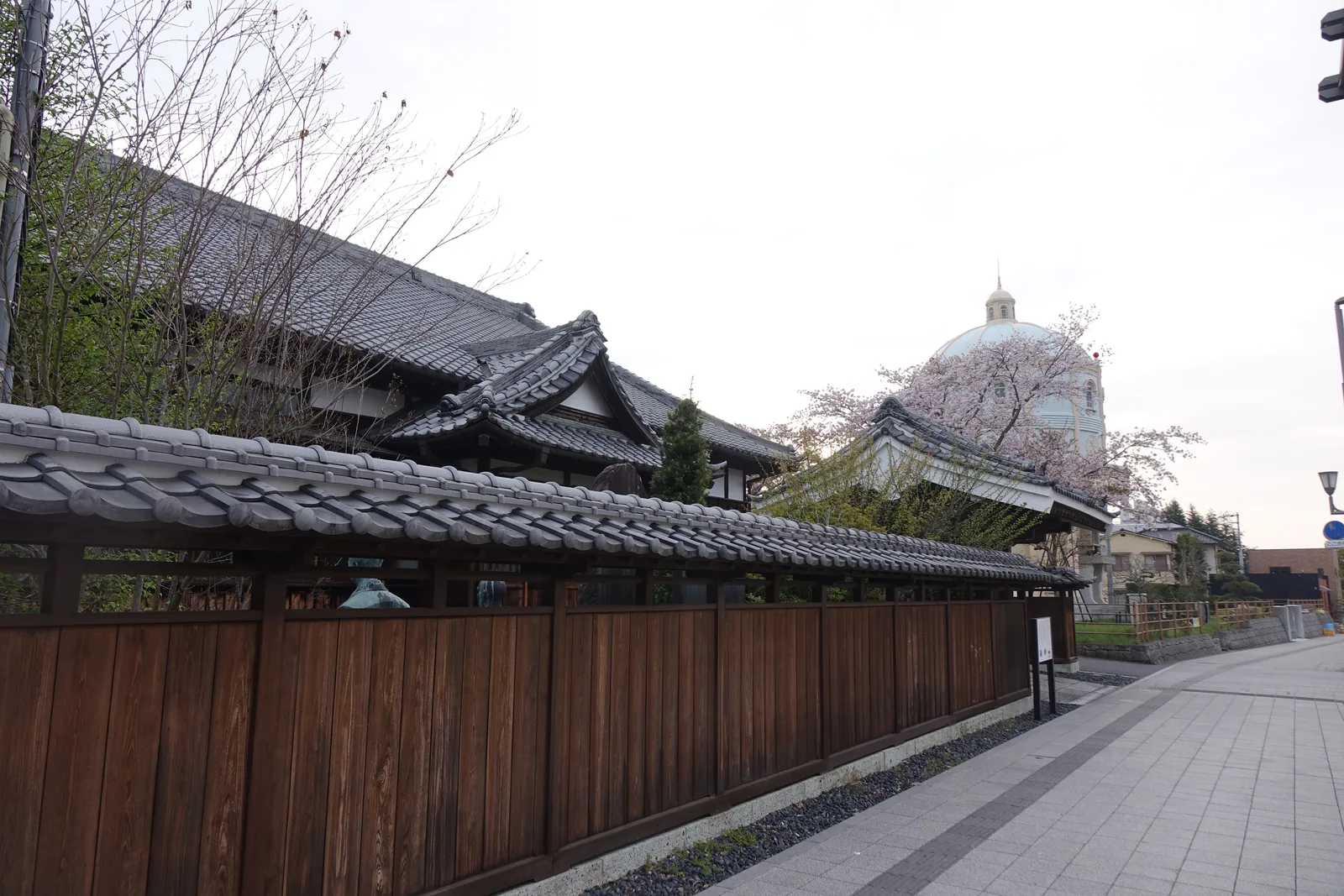 沿人行道延伸的深色木板塀，塀頂覆瓦，板塀後方露出建築的瓦屋頂與遠處的配水塔圓頂