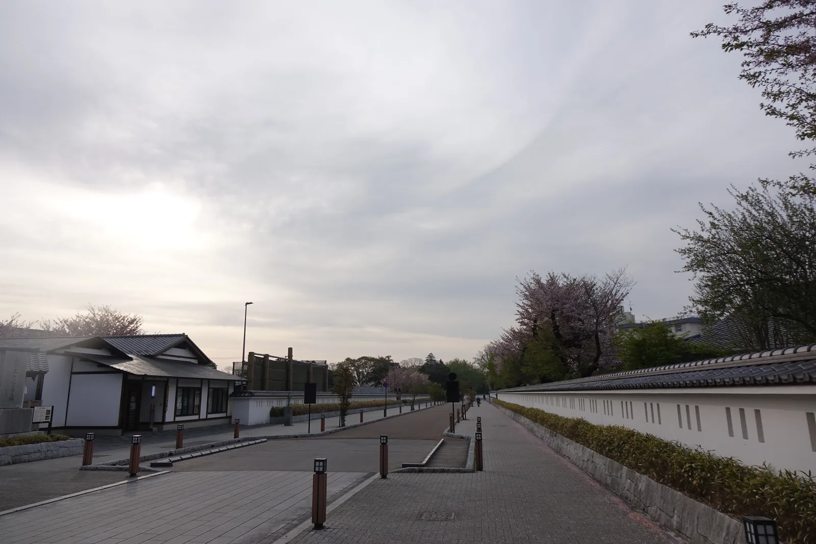 二之丸的直線道路，右側是白色的土塀，塀上開有縱長的小窗，左側是低矮的車擋與植栽，道路盡頭是校舍與櫻樹