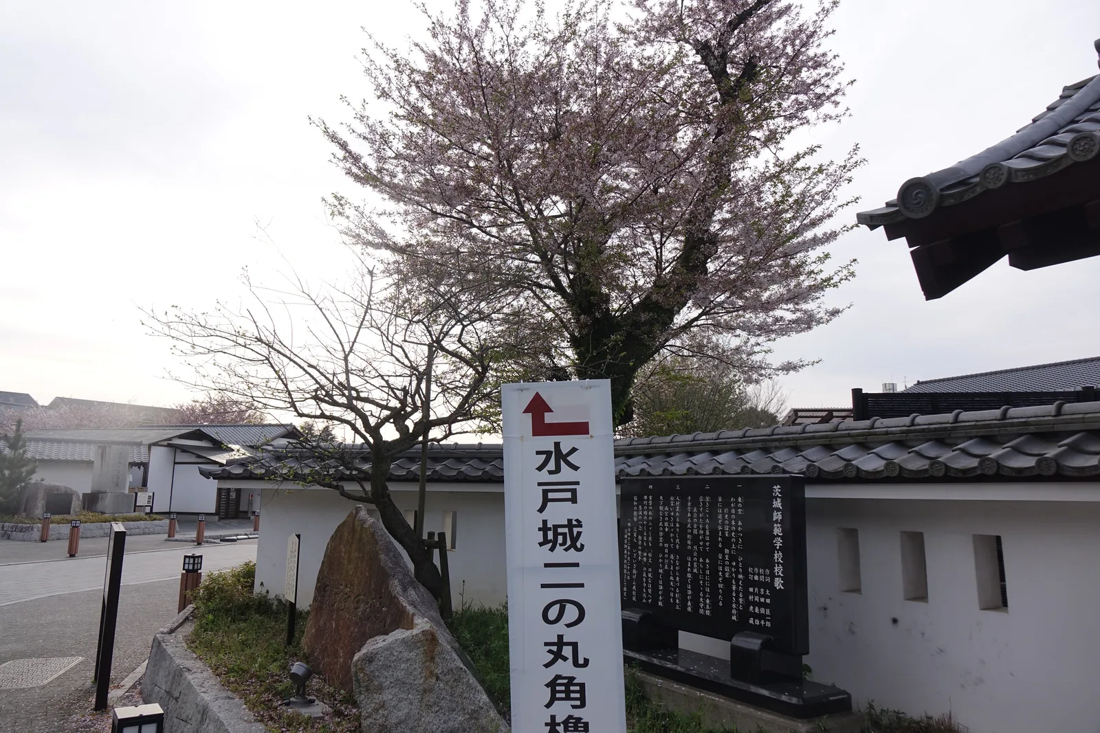 白色土塀的端部與一根紅色的直書路標，路標上寫著水戶城二之丸角櫓，旁邊有刻字的石碑與覆瓦的塀頂，遠處是道路與校舍