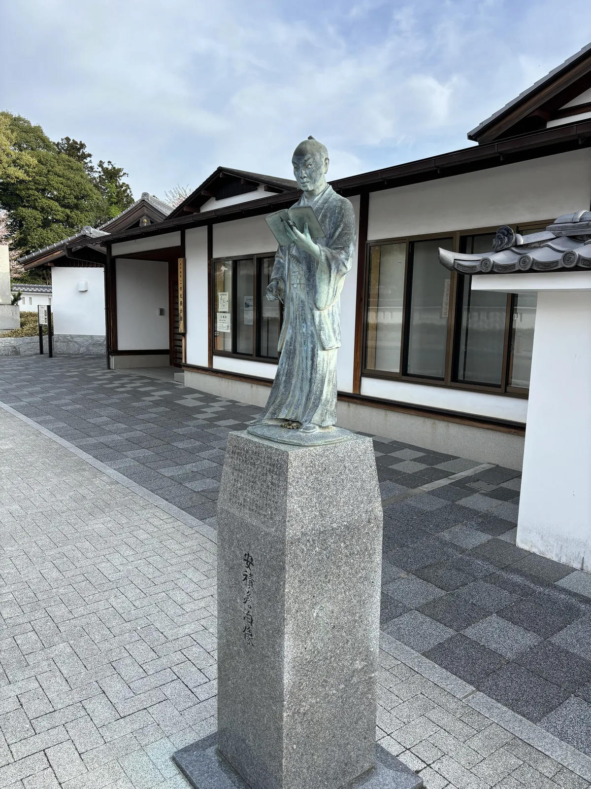 展示館前的銅像，一位著長袍、手持書卷的人物立在灰色花崗岩基座上，背後是白牆與深色出簷的低層建築，地面鋪著深淺兩色的方磚