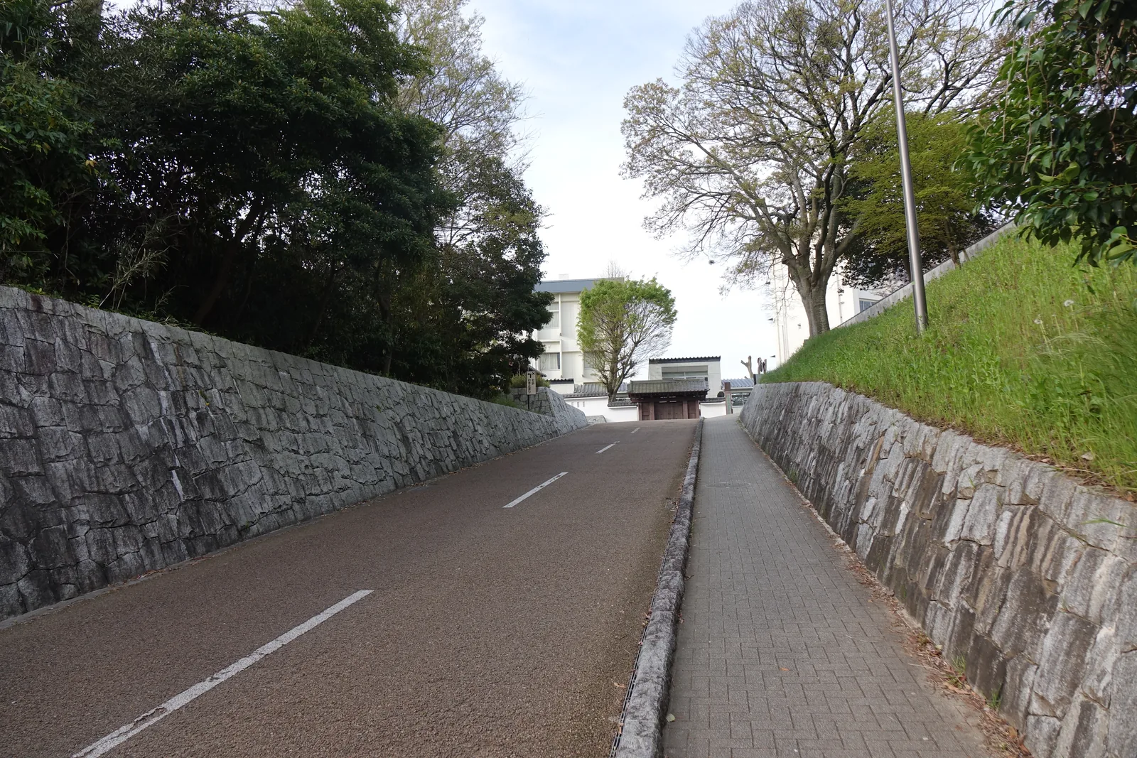 兩道石垣之間的下坡道路，右側是長滿草的土坡與樹木，道路盡頭可見白色的建築與覆瓦的門