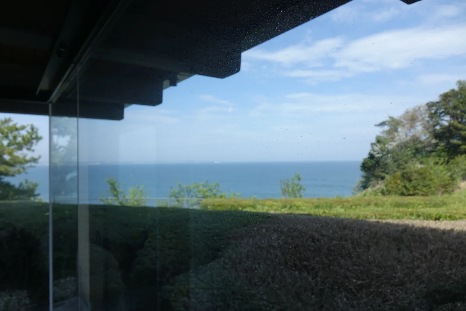 自館內望向海面，畫面上方是深色的簷口與階梯狀的梁端，中段是一片玻璃，玻璃外是一片低矮的綠籬與草坡，遠方是藍色的太平洋與水平線