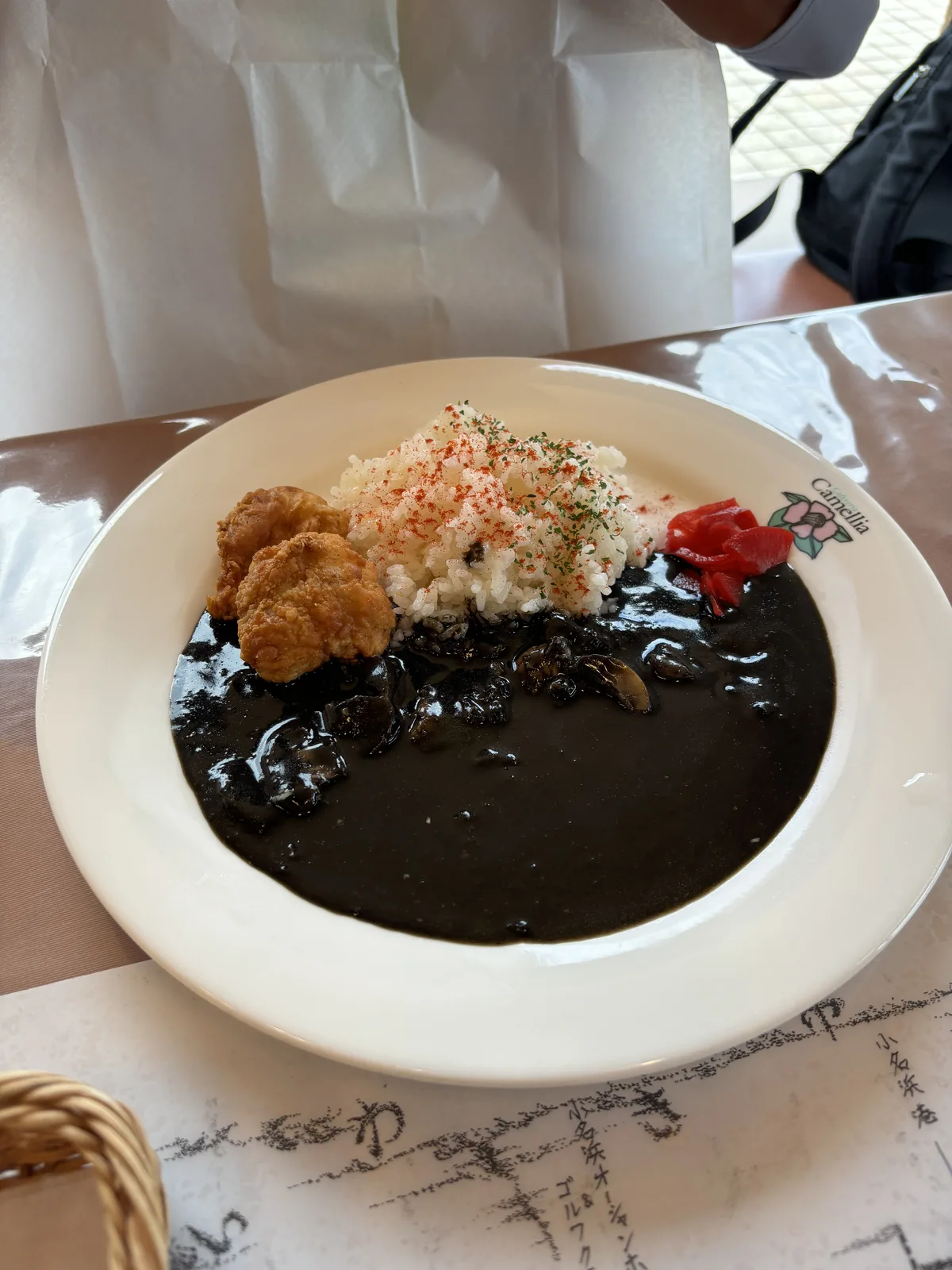 白色餐盤上盛著黑色的咖哩、米飯與炸物，飯上撒著配料，旁邊放著紅色的醃菜，餐盤下方墊著印有素描圖案的紙墊