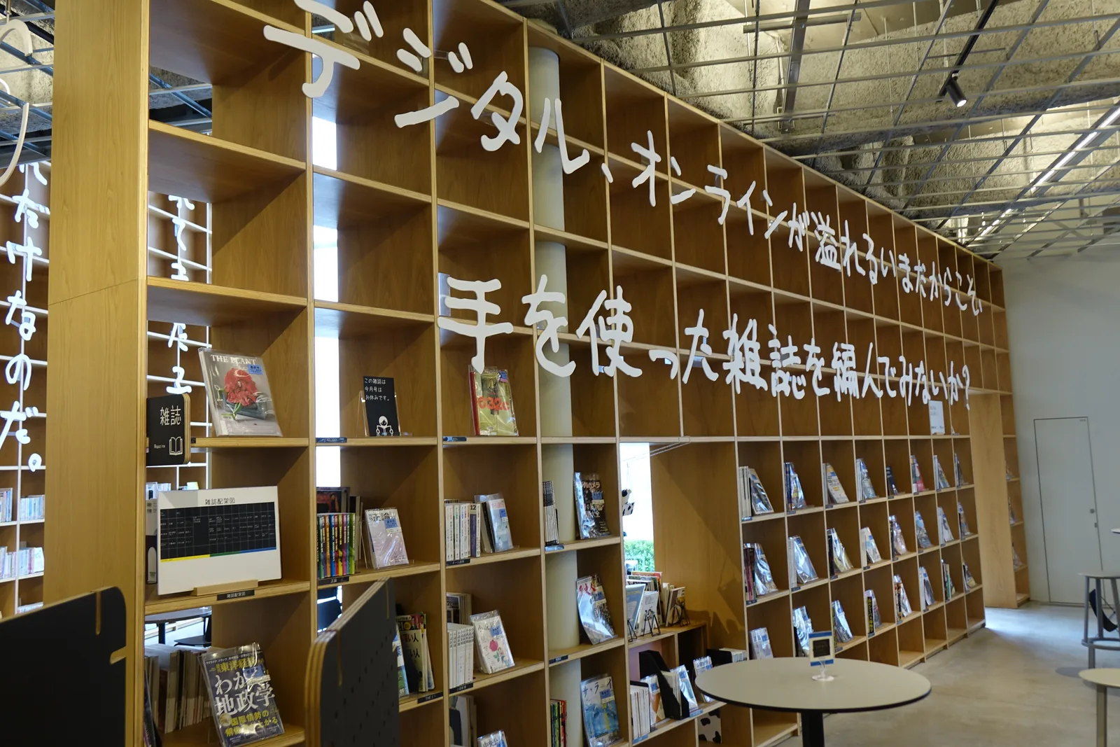 一樓的木格書架上方，以白色的大字手寫體寫著兩行句子，書架中陳列雜誌，前方放著一張圓桌